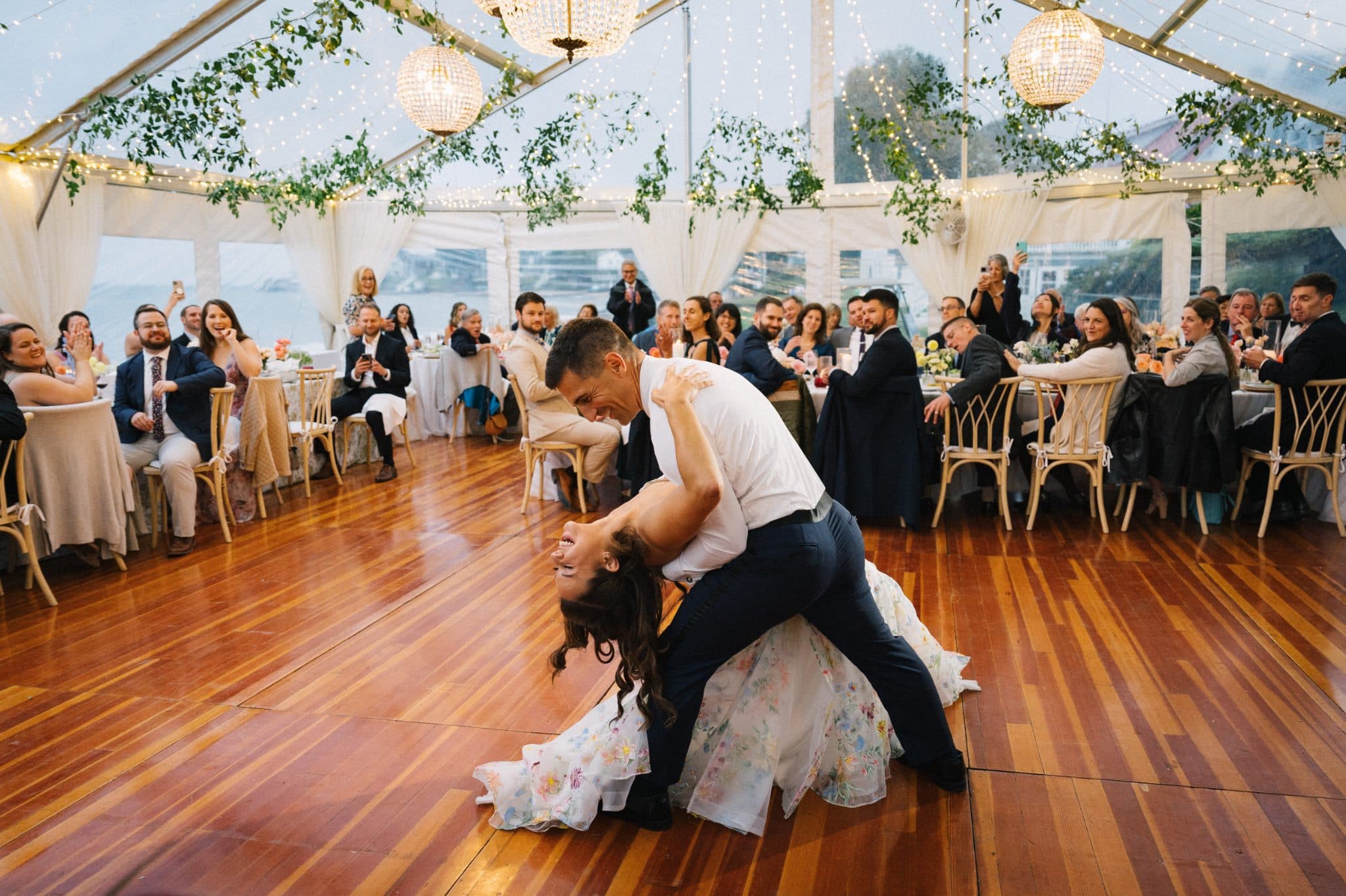 A couple dances on a wooden floor in a tent with guests seated at tables in the background.