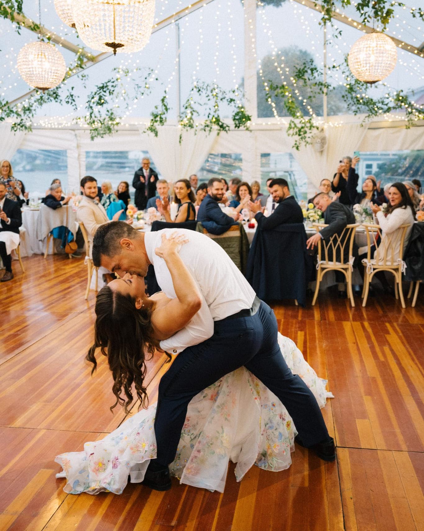 A couple dances closely on a wooden floor under a tent adorned with lights and greenery.
