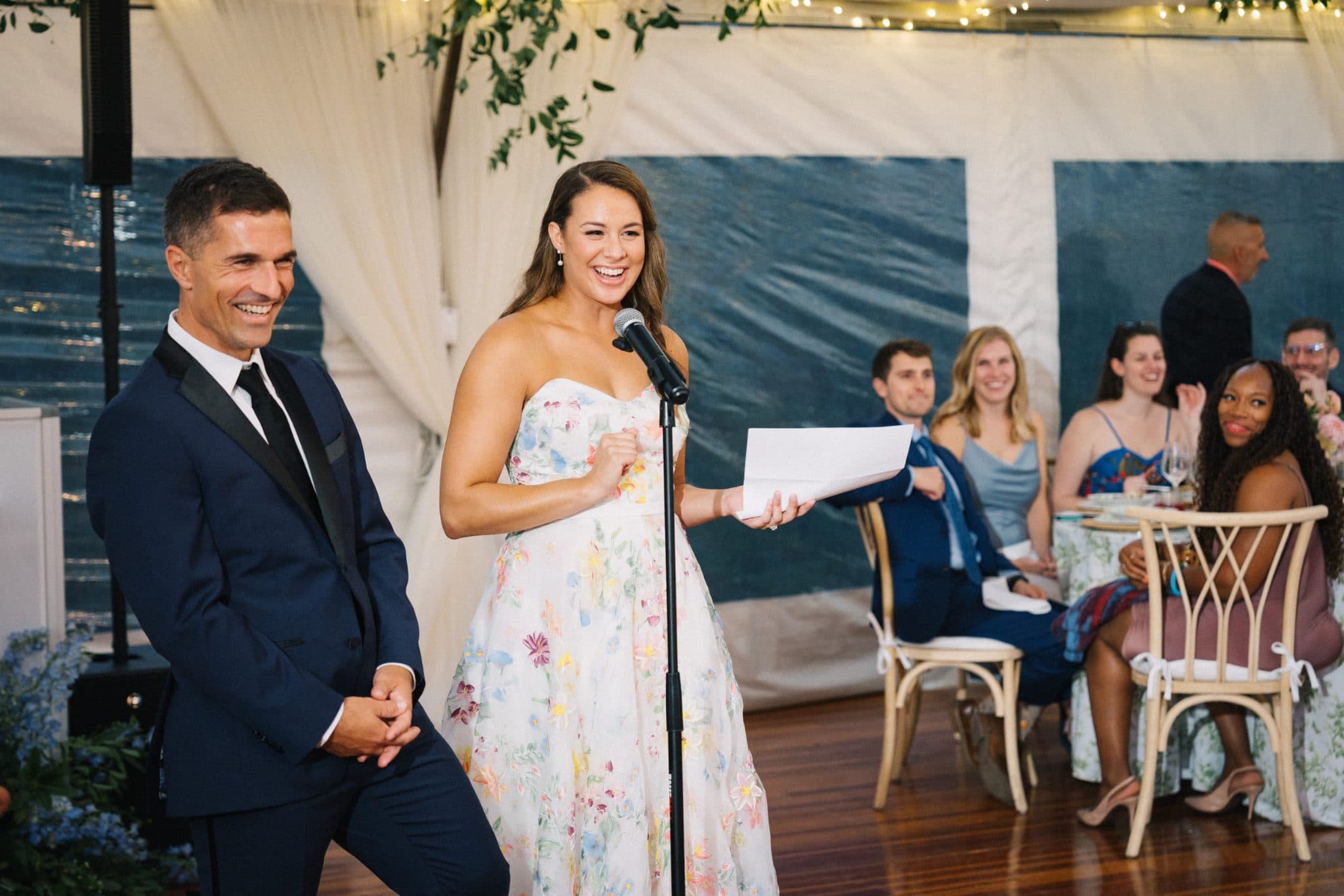 A woman in a floral dress stands at a microphone, smiling, while a man in a suit stands beside her, both in a tented wedding setting with guests seated in the background.