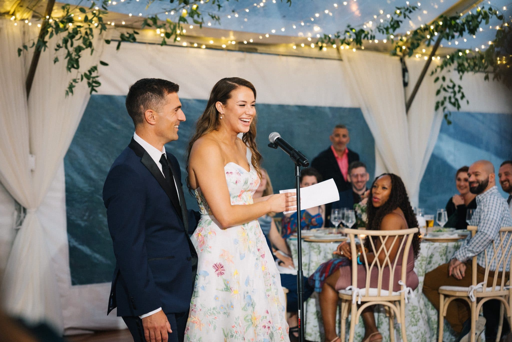 A woman in a floral dress stands at a microphone, smiling, with a man in a suit beside her, while guests sit at tables in the background.