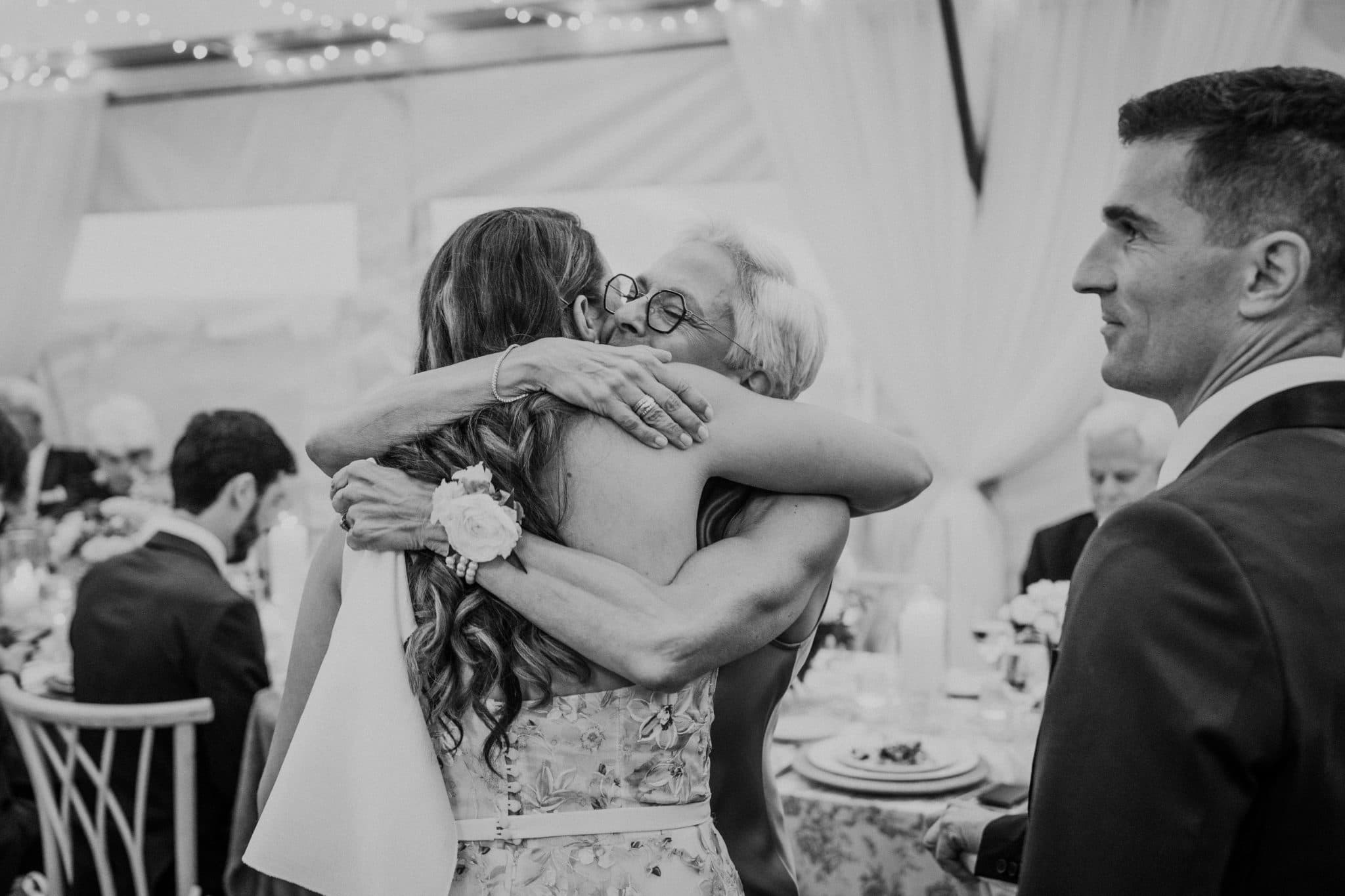 A bride hugs her father at a wedding reception, with guests seated at tables in the background.