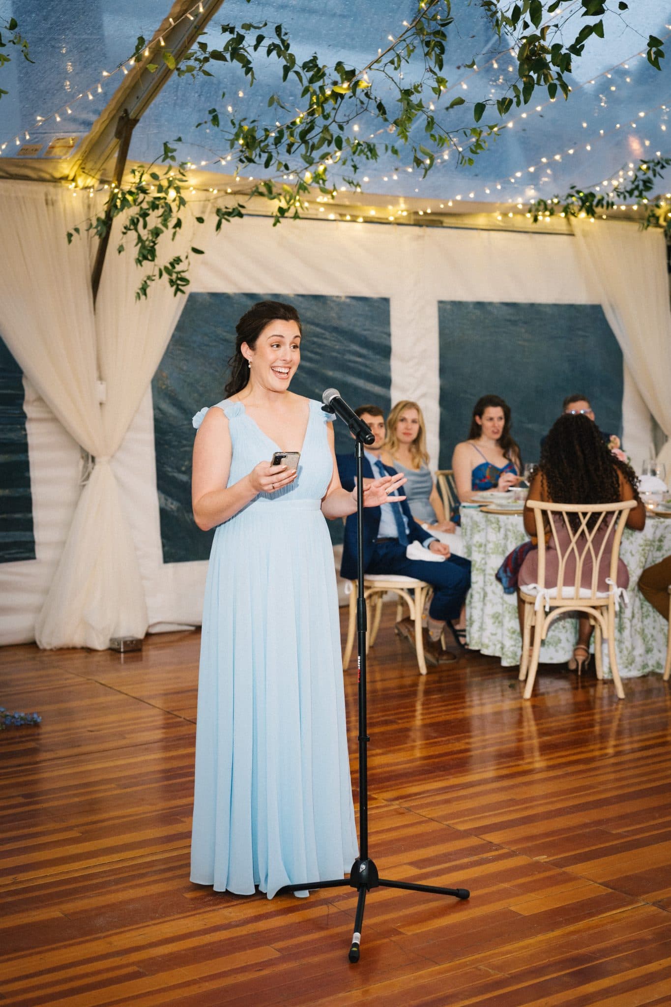A woman in a light blue dress stands at a microphone, smiling, with a group of seated guests in the background under a tent decorated with lights and greenery.