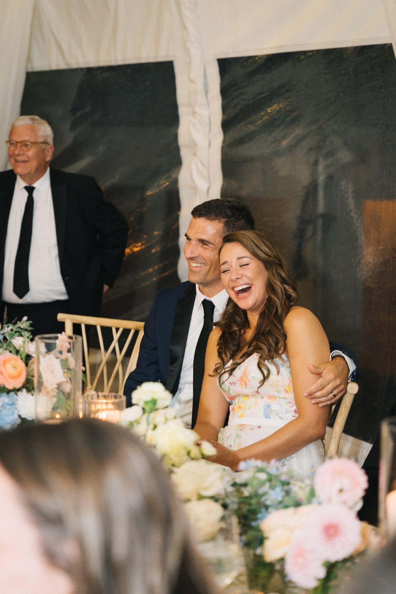 A couple sits at a table adorned with flowers, smiling and laughing during a wedding reception.