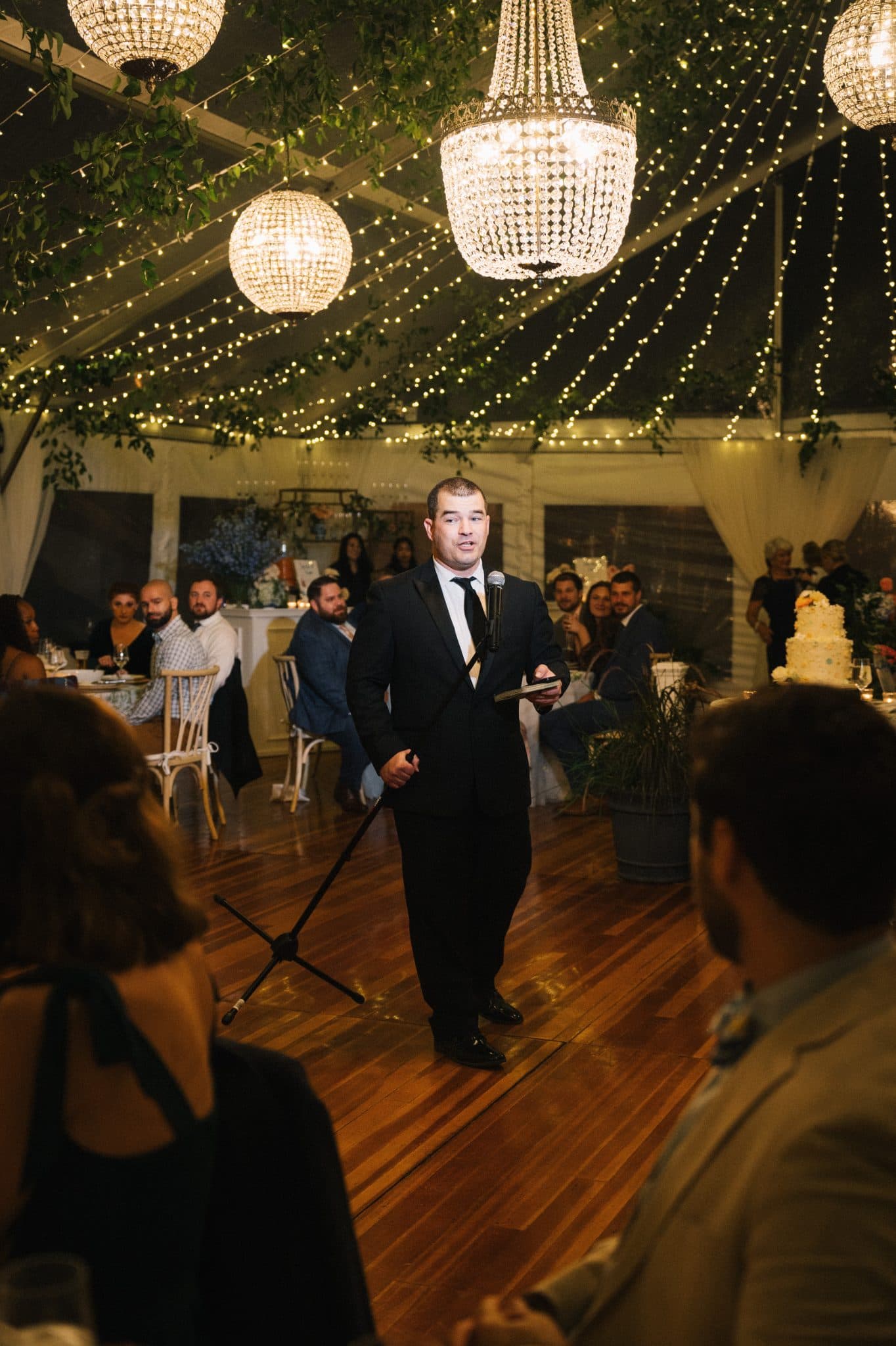 A man in a suit stands on a wooden floor holding a microphone, addressing a seated audience under a canopy of lights and chandeliers.