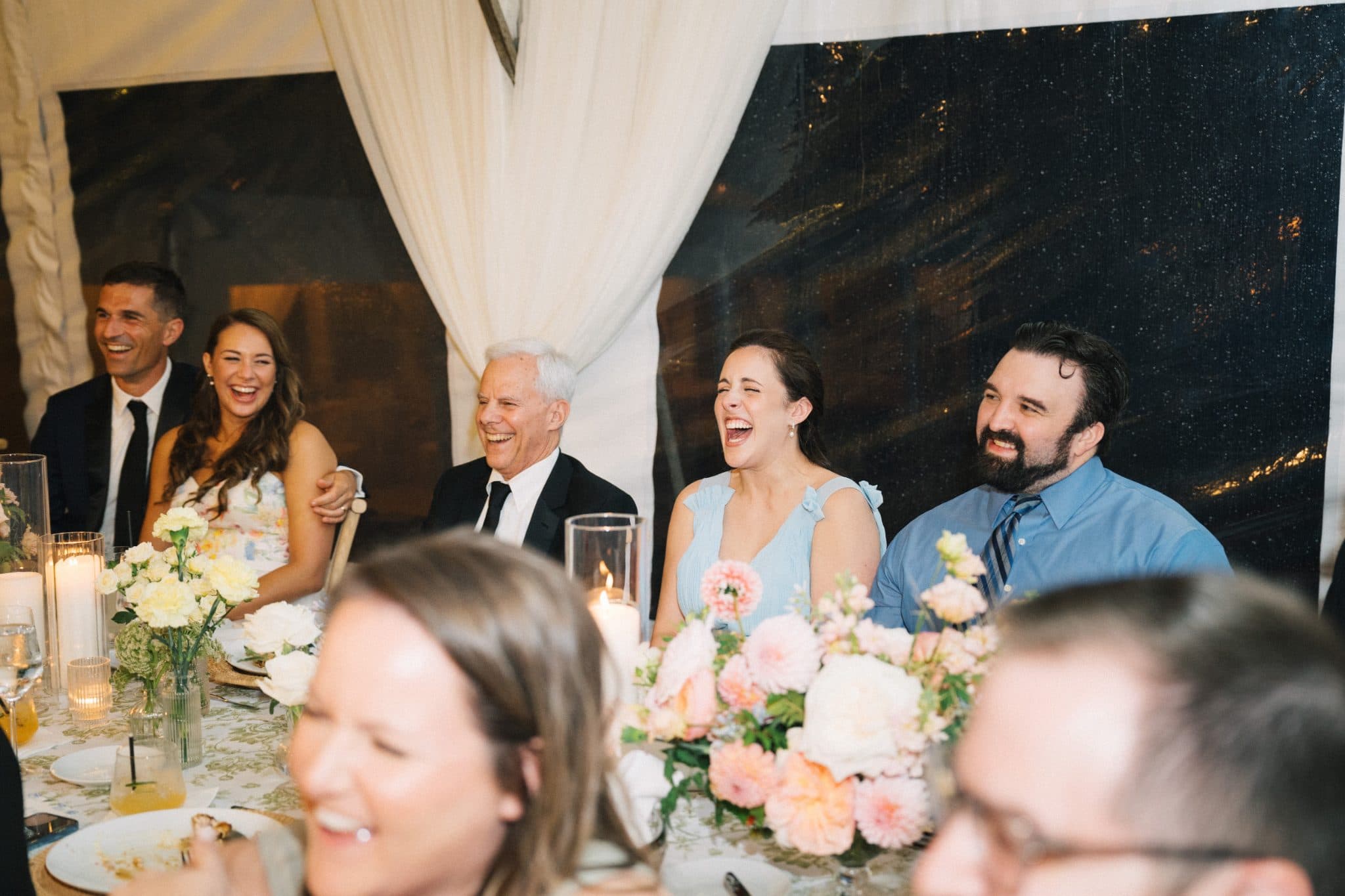 A group of people sits at a table adorned with flowers and candles, laughing and enjoying a festive moment.