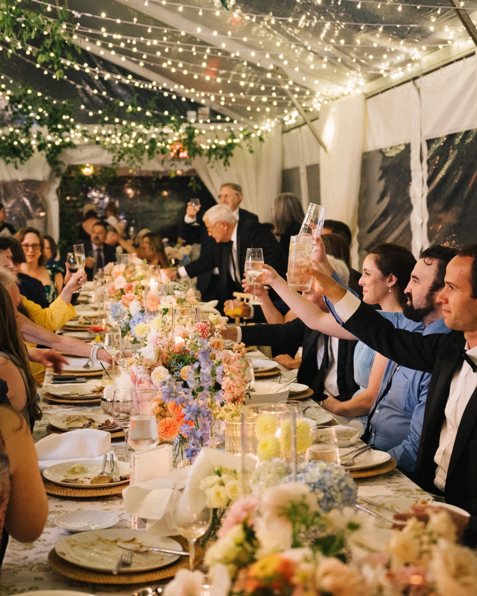 Guests at a wedding reception raise their glasses in a toast under string lights and floral decorations.