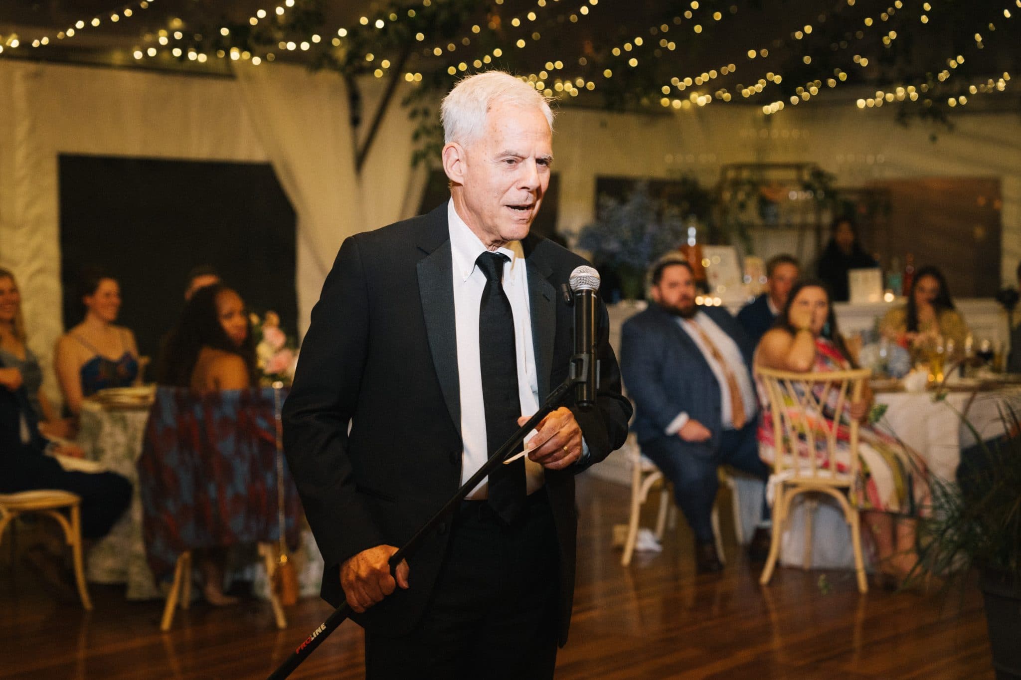 A man in a suit holds a cane and a microphone, speaking at a wedding reception with guests seated at tables.