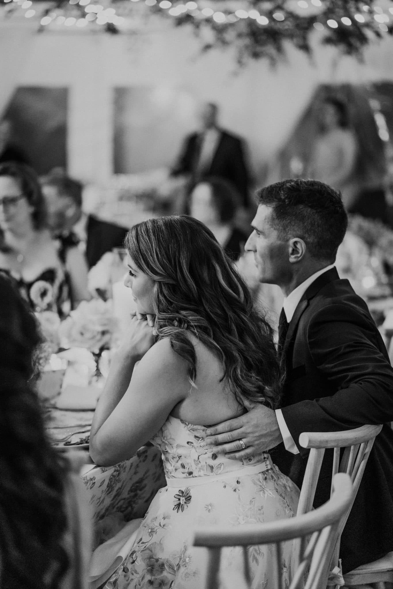 A bride in a floral dress sits with her arm around a man in a suit at a wedding reception.