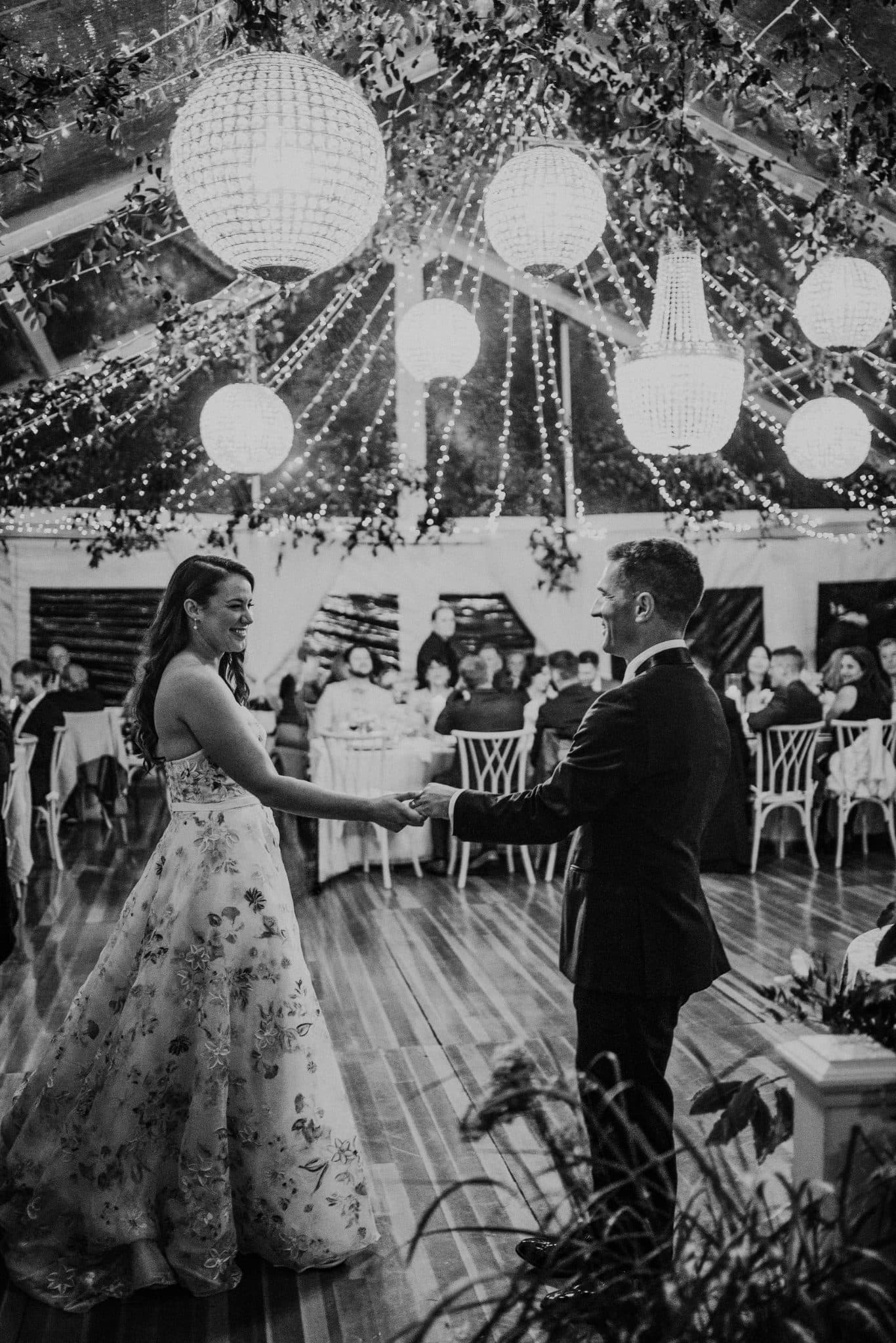 A bride in a floral gown and a groom in a suit dance under a canopy of lights and greenery.