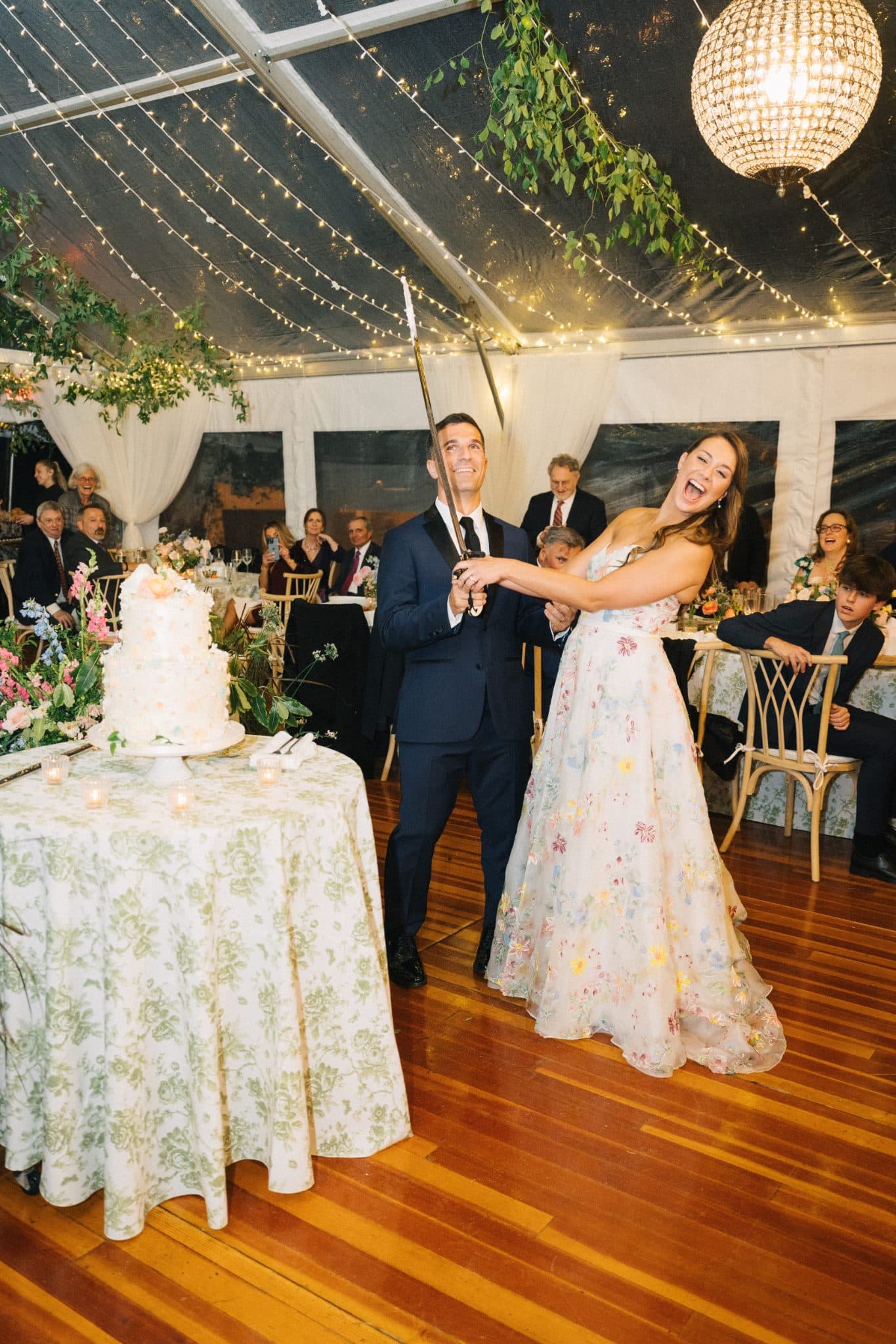 A bride and groom celebrate at their wedding reception, cutting a cake under a tent decorated with fairy lights and greenery.