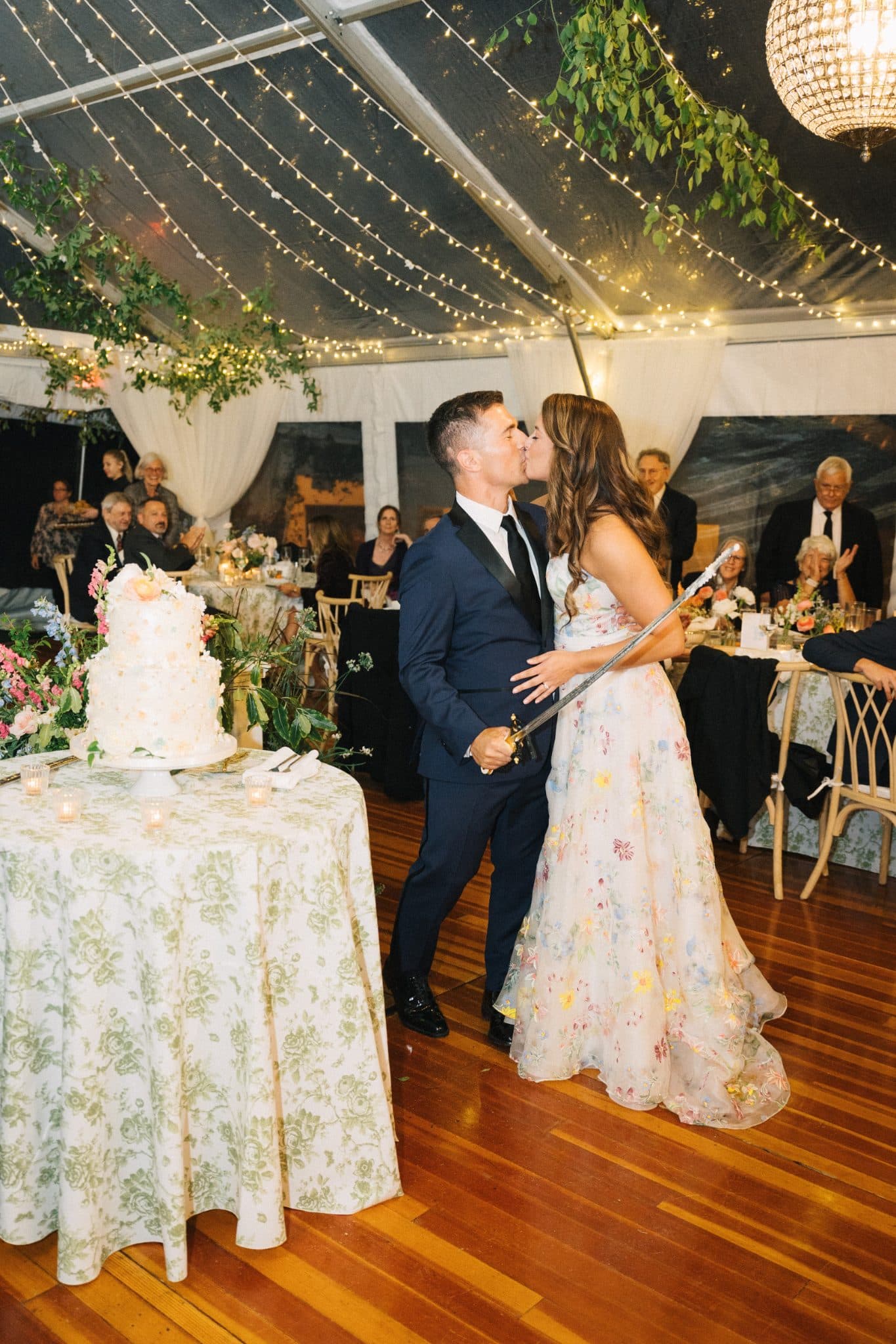 A couple kisses near a wedding cake at a tented reception with string lights and floral decorations.