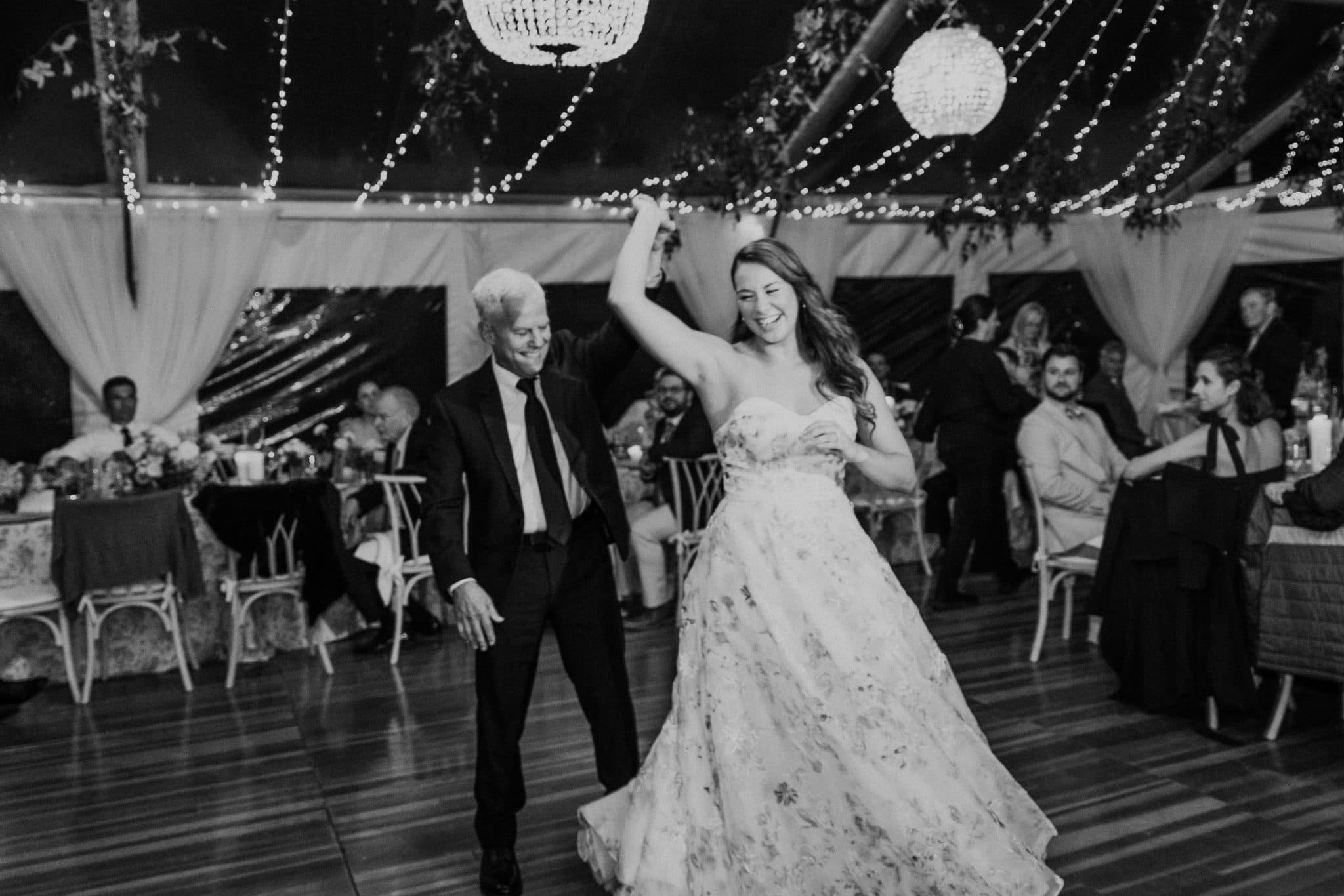 A bride in a strapless gown dances with a man in a suit under a tent adorned with lights.