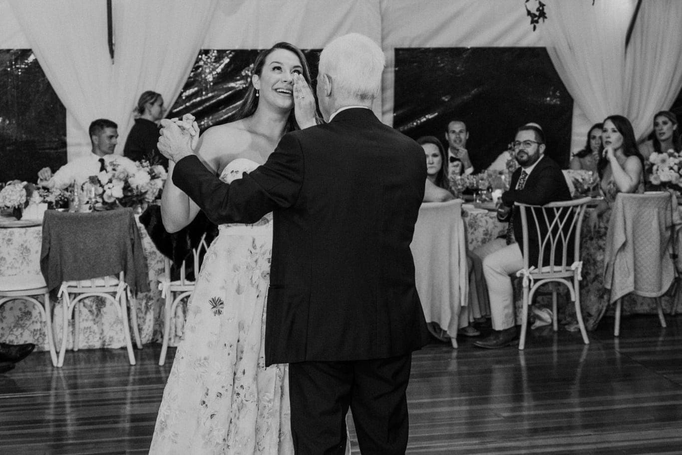 A bride and groom dance under chandeliers and string lights at a wedding reception.