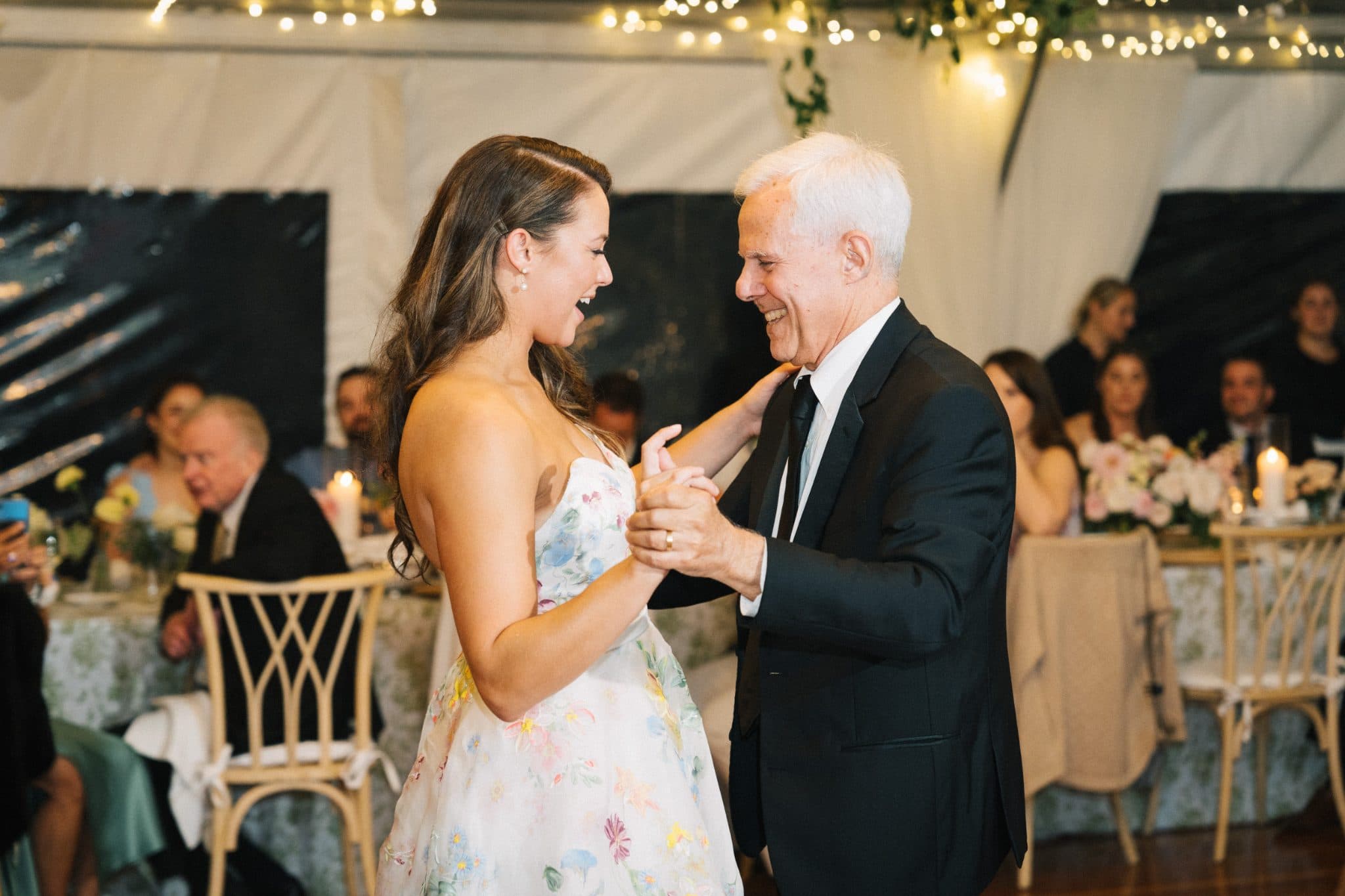 A woman in a floral dress dances with a man in a suit under twinkling lights.