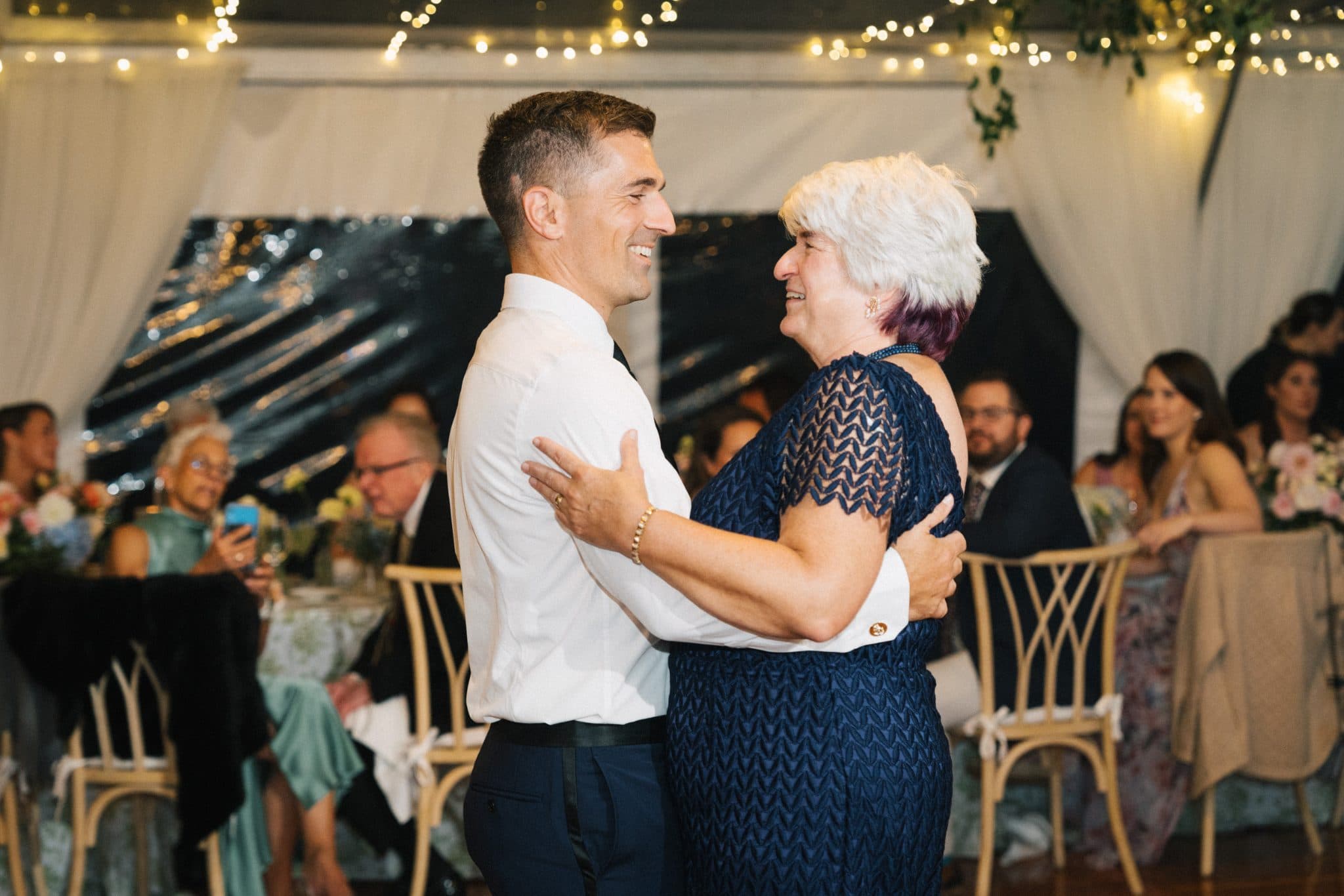 A man and woman dance closely, smiling, in a warmly lit wedding reception setting.