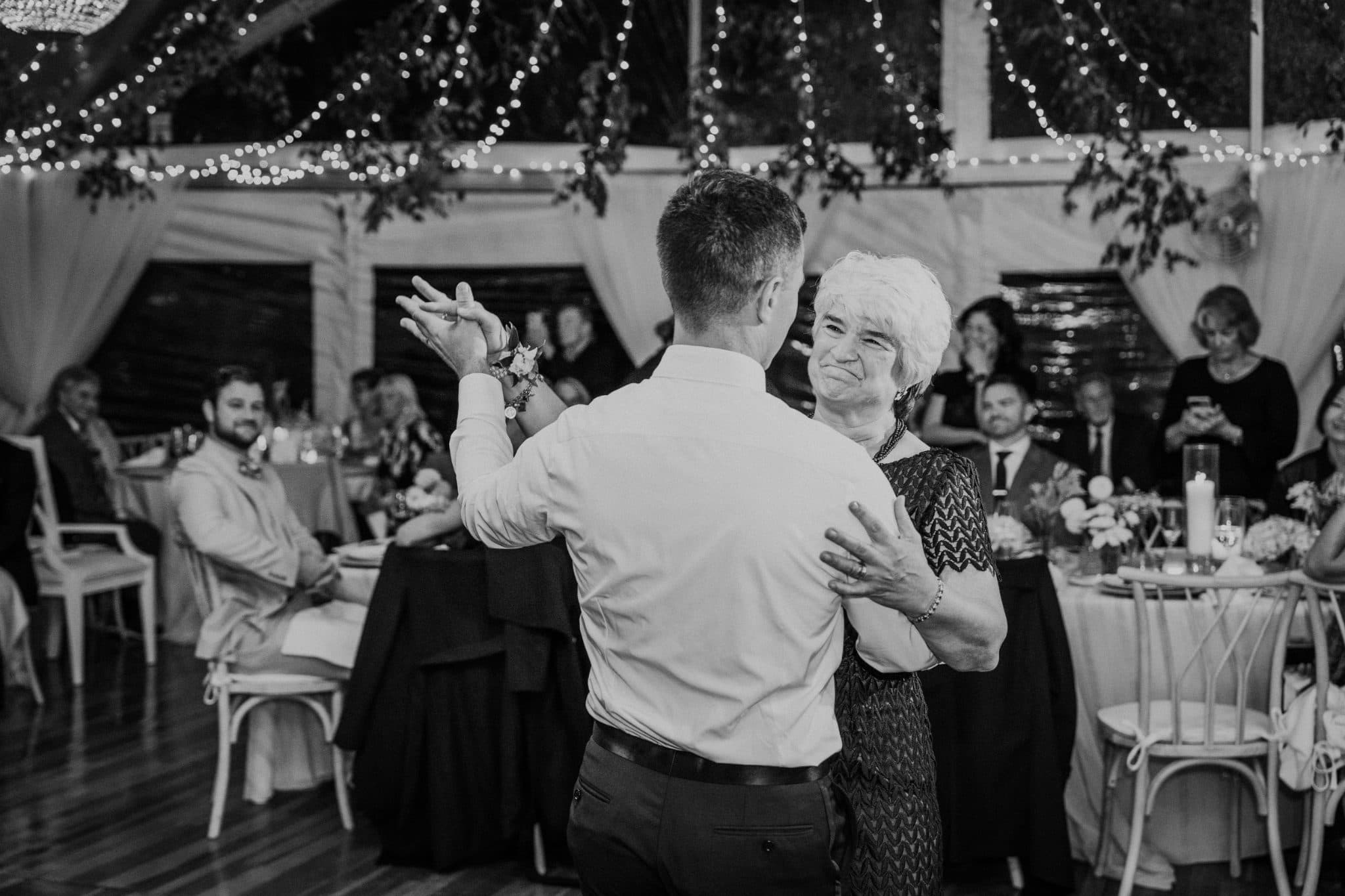A man and woman dance together on a wooden floor under string lights, surrounded by seated guests at a wedding reception.