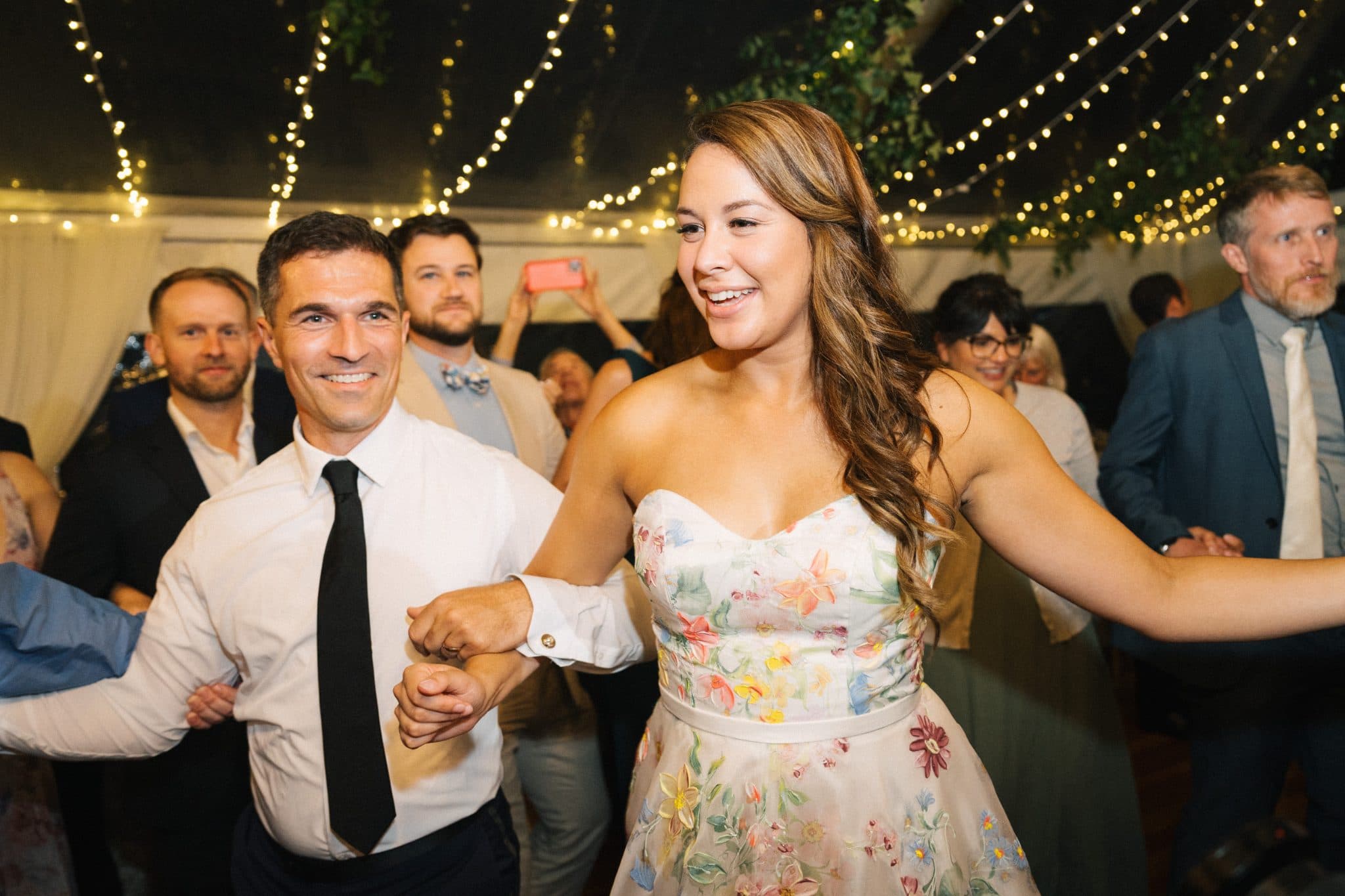 A woman in a floral dress dances with a man in a white shirt and black tie at a wedding reception under string lights.
