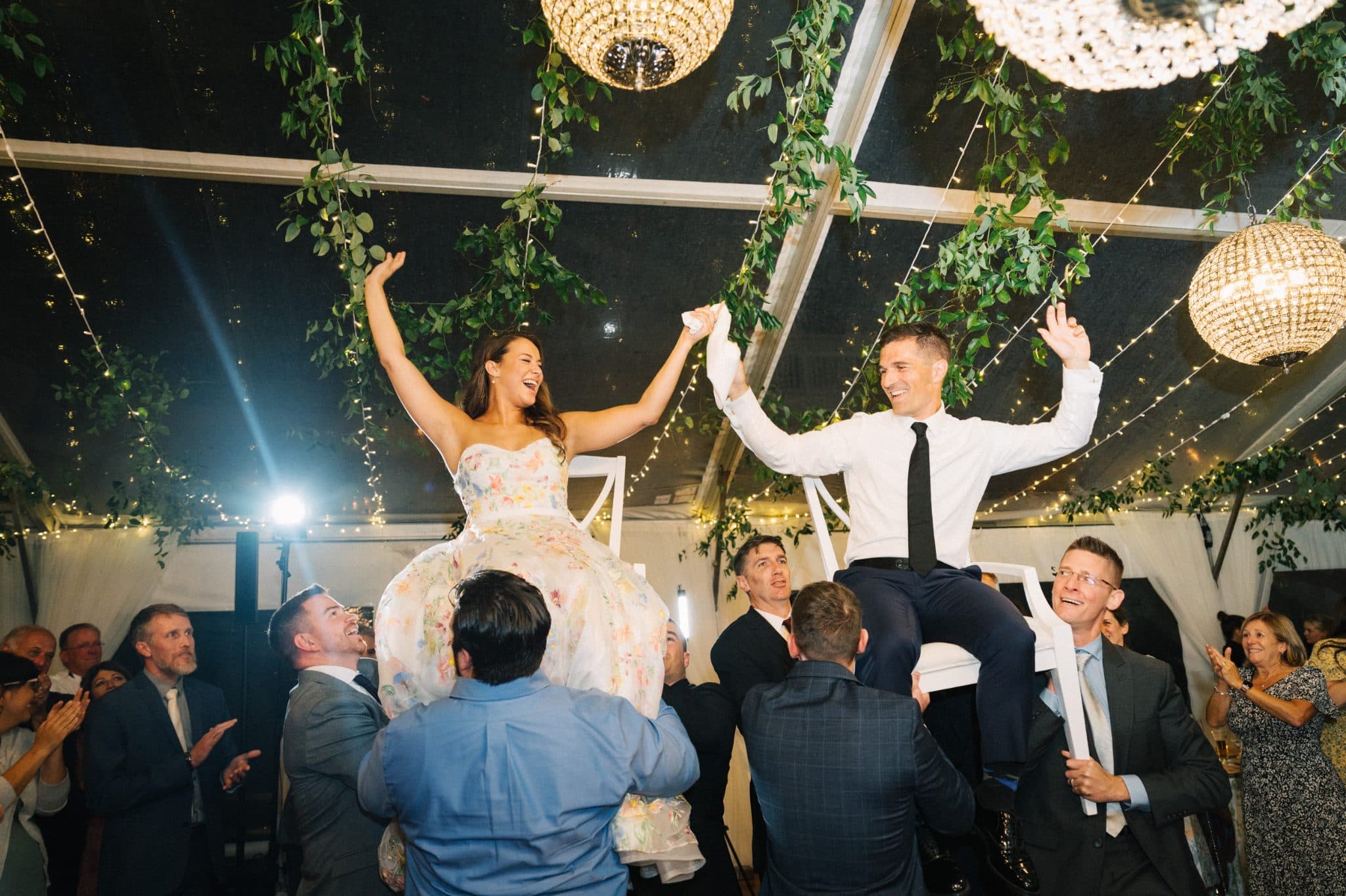 A bride and groom are seated on chairs, held aloft by their wedding guests in a decorated tent with greenery and lights.