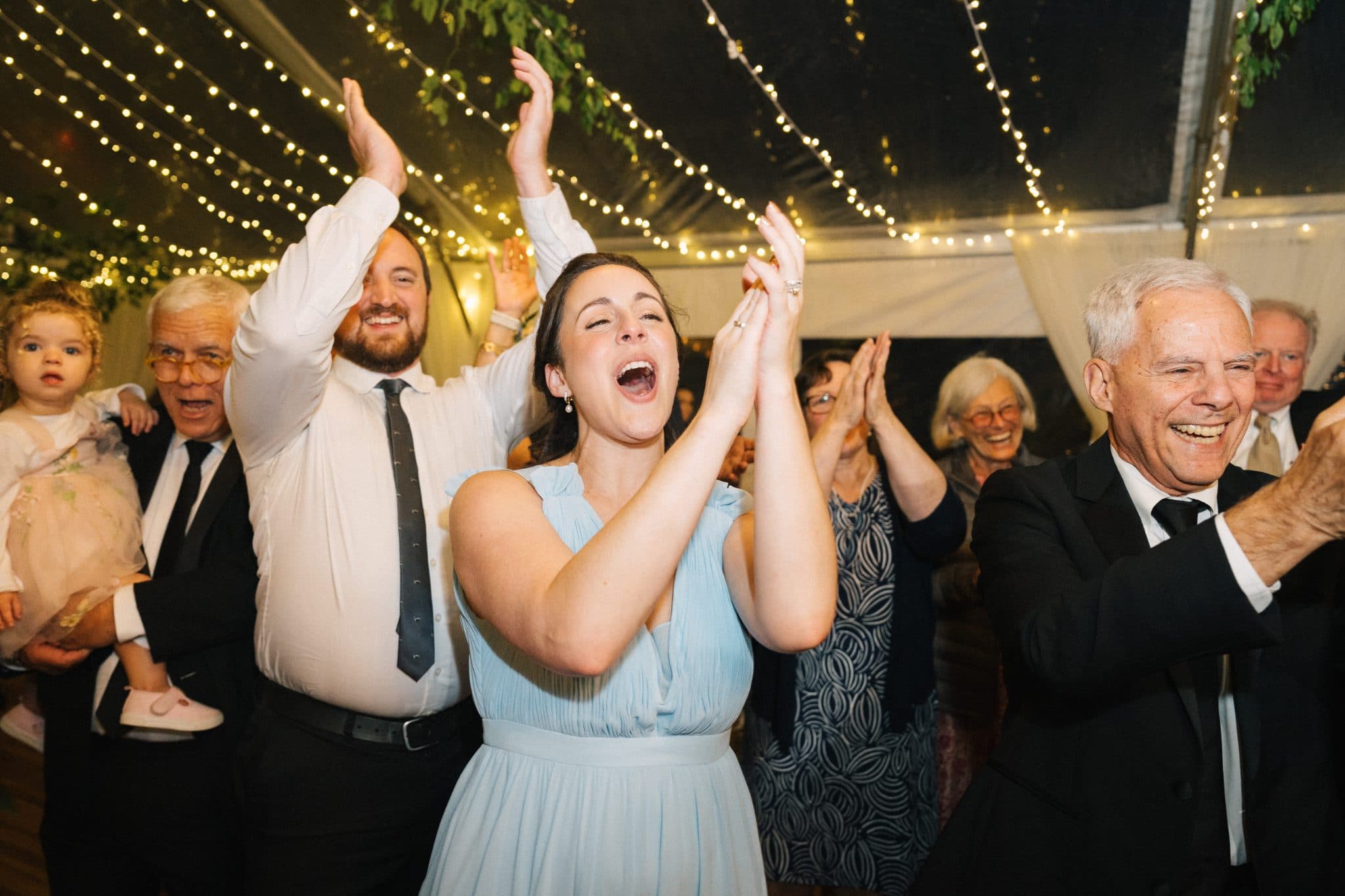 A woman in a light blue dress claps enthusiastically, surrounded by guests in formal attire under string lights.