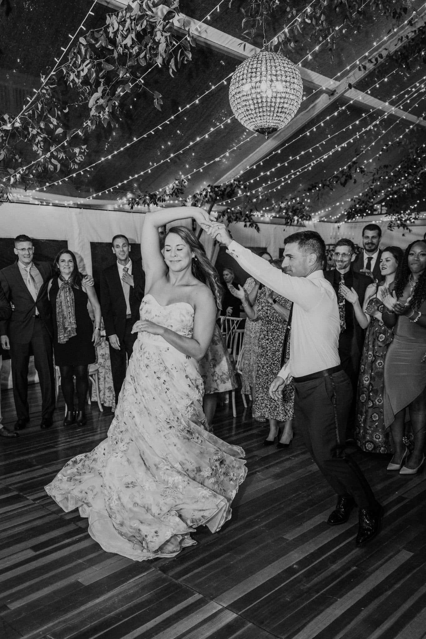 A bride in a floral gown dances with a man in a white shirt and dark pants under a disco ball and string lights.