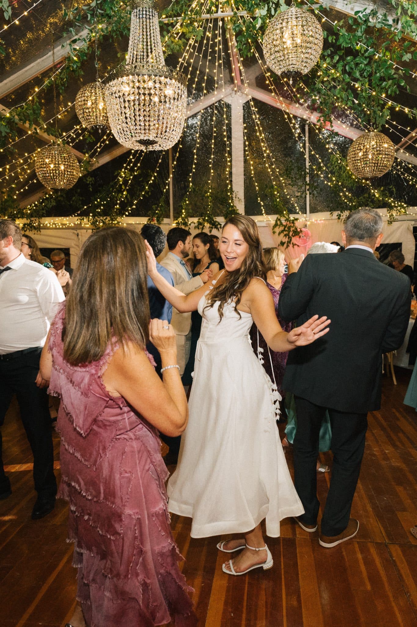 A bride dances joyfully with guests under a canopy of lights and greenery.