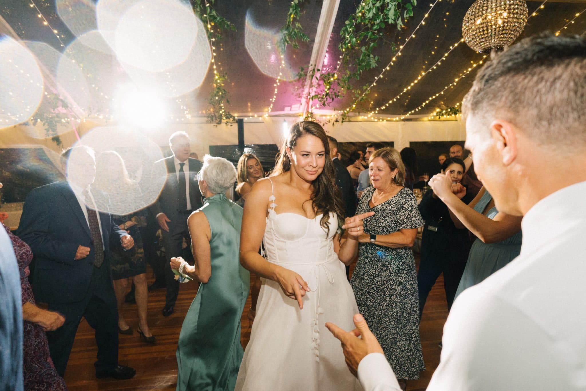 A bride in a white dress dances surrounded by guests in a warmly lit, decorated tent.
