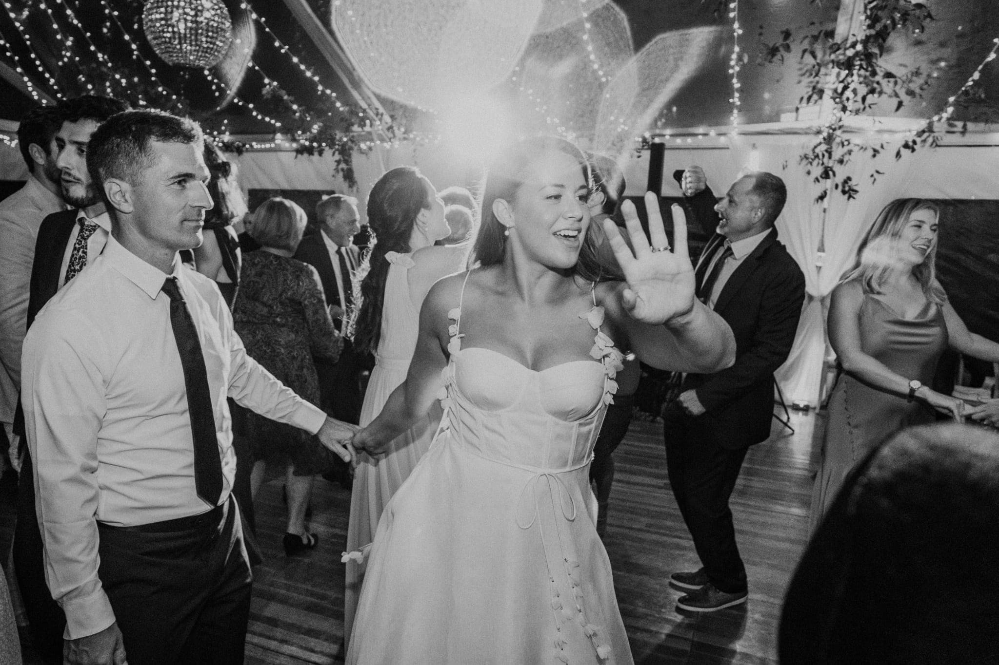 A bride in a strapless dress dances with a man in a white shirt and tie, surrounded by guests in a tent with string lights and disco balls.