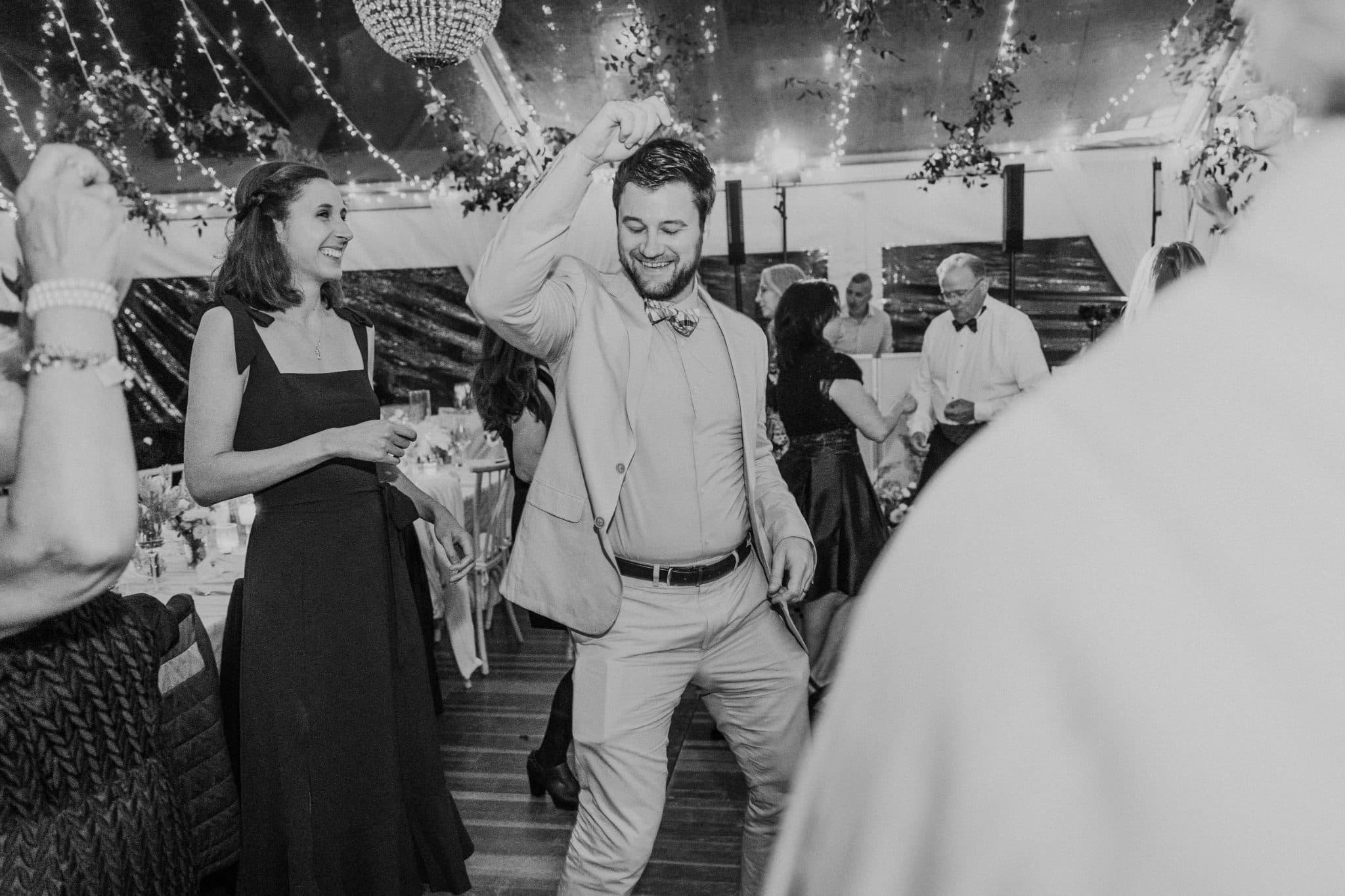 A man in a suit dances joyfully with a woman in a black dress under a tent adorned with lights and flowers.