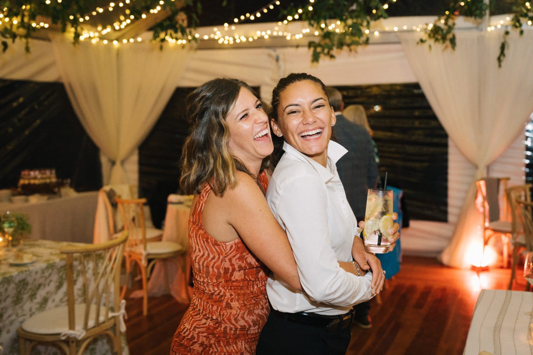 Two women are embracing and smiling at a wedding reception with string lights and tables in the background.