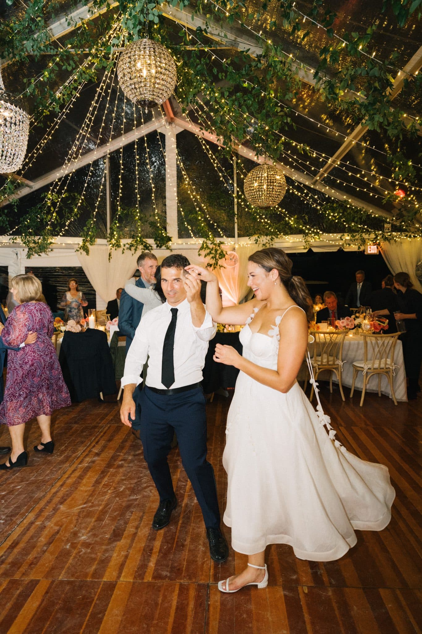 A bride and groom dance joyfully on a wooden floor under a canopy of greenery and twinkling lights.