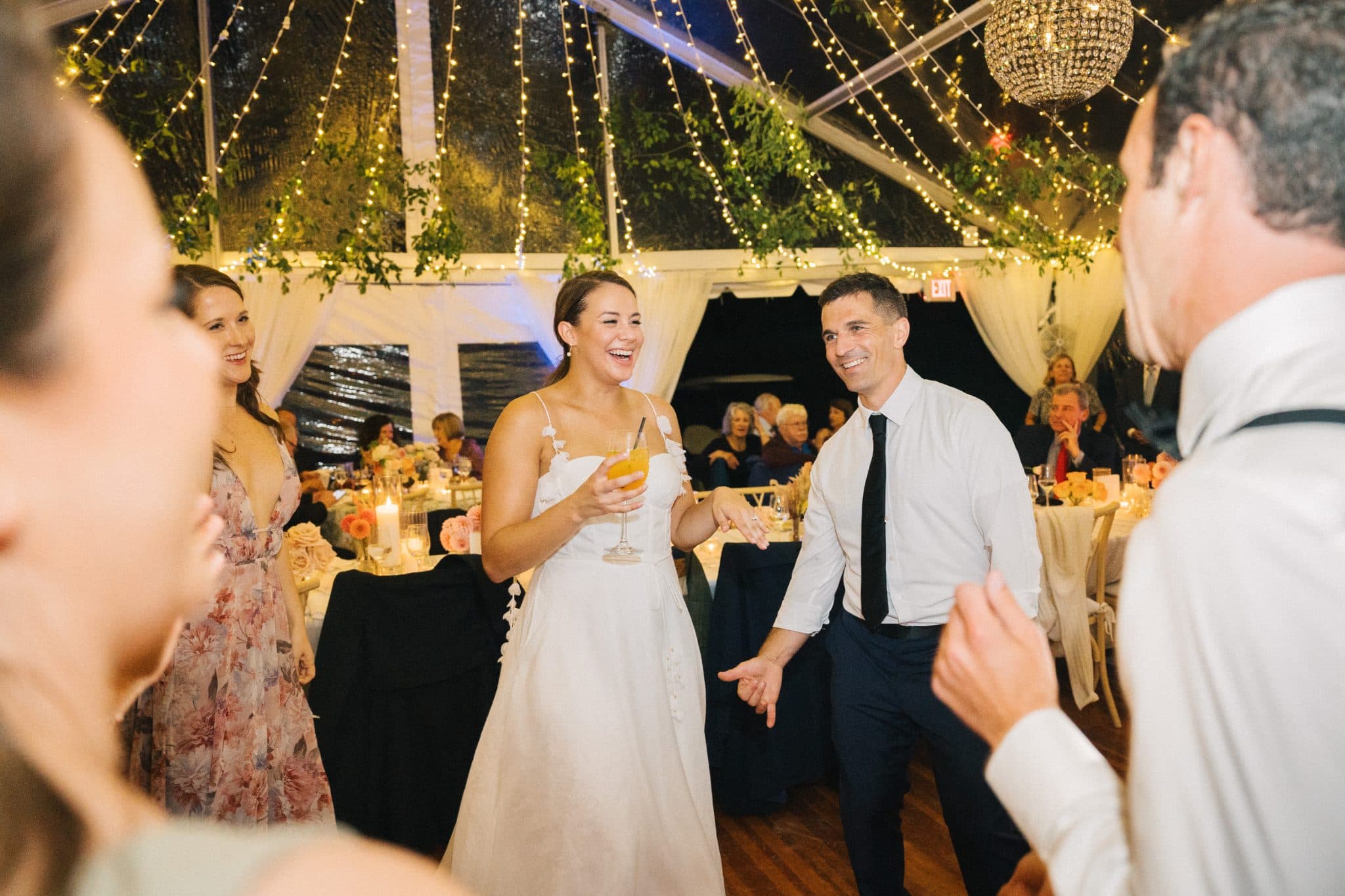 A bride in a white dress dances with a man in a white shirt and black tie under a tent adorned with lights and greenery.
