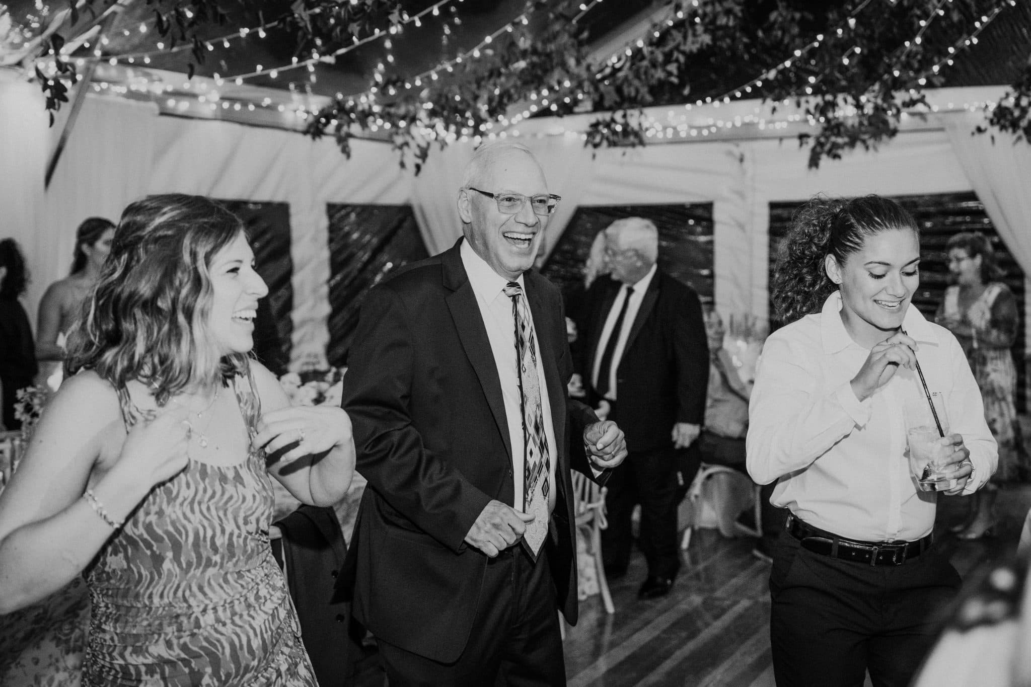 A black-and-white wedding photograph shows three people dancing joyfully under a tent decorated with string lights.