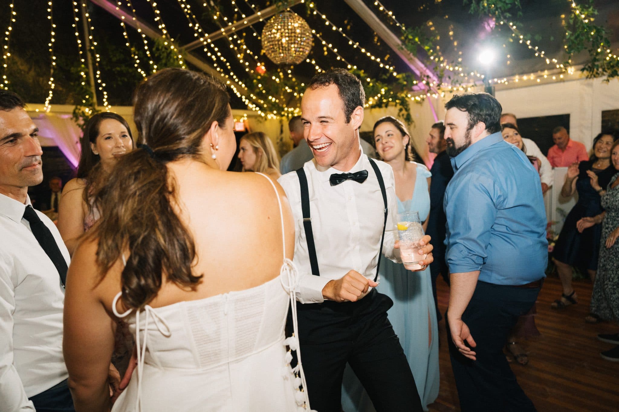 A bride and groom dance joyfully at a wedding reception under string lights and a disco ball.