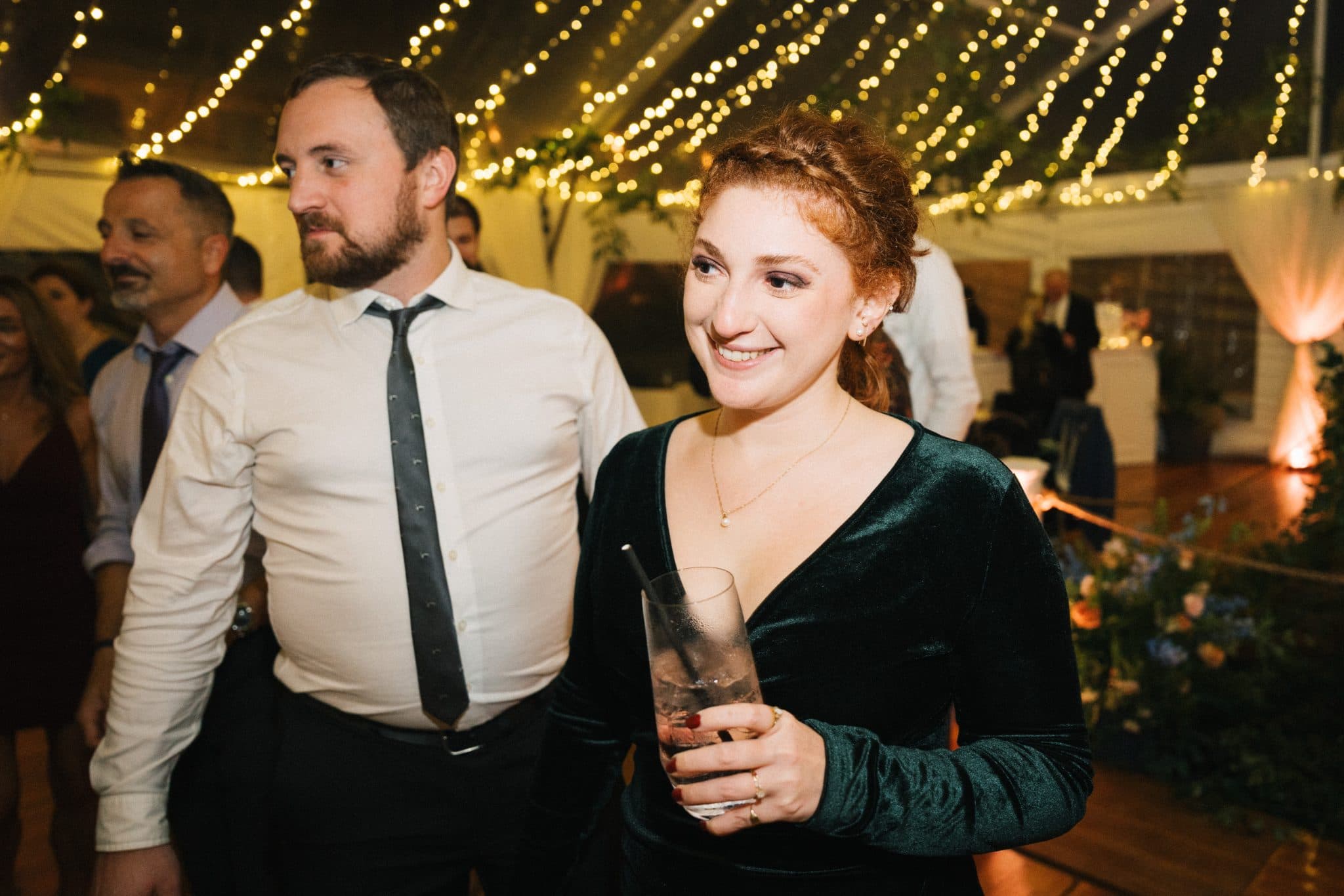 A woman in a green velvet dress holds a drink, smiling, while a man in a white shirt and tie stands beside her at a festive event.