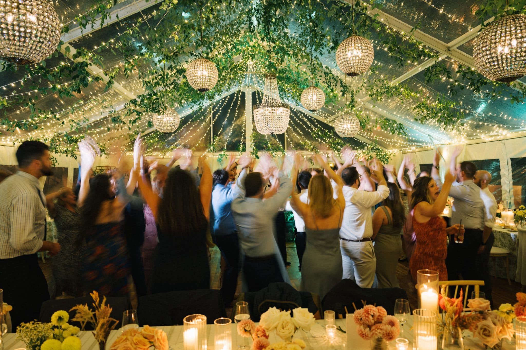 A lively wedding reception with guests raising their hands, adorned with greenery, string lights, and elegant chandeliers.