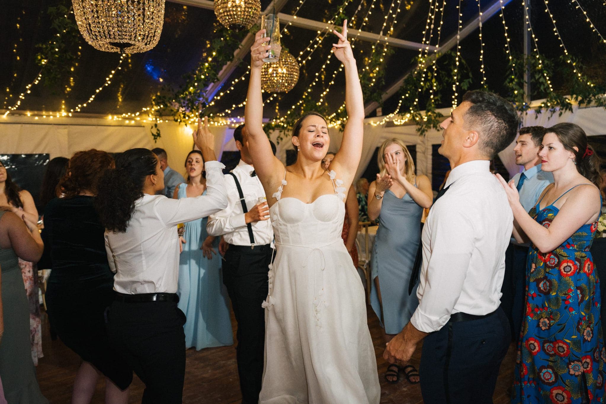 A bride in a white dress dances joyfully with guests under a tent adorned with string lights and chandeliers.