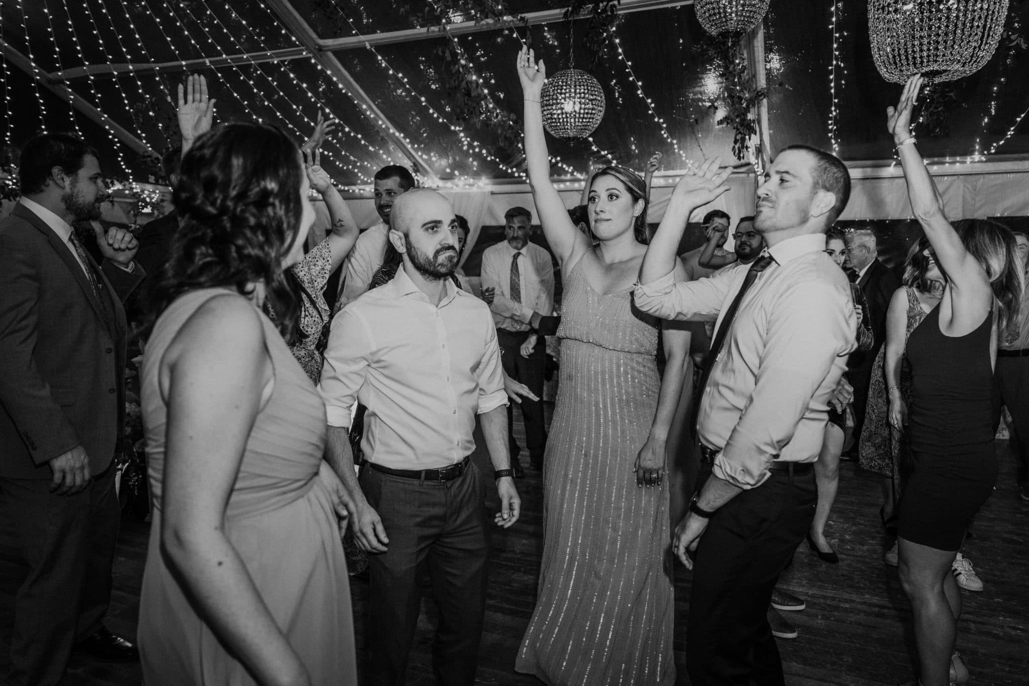 A group of people dances joyfully under a tent adorned with string lights and chandeliers.