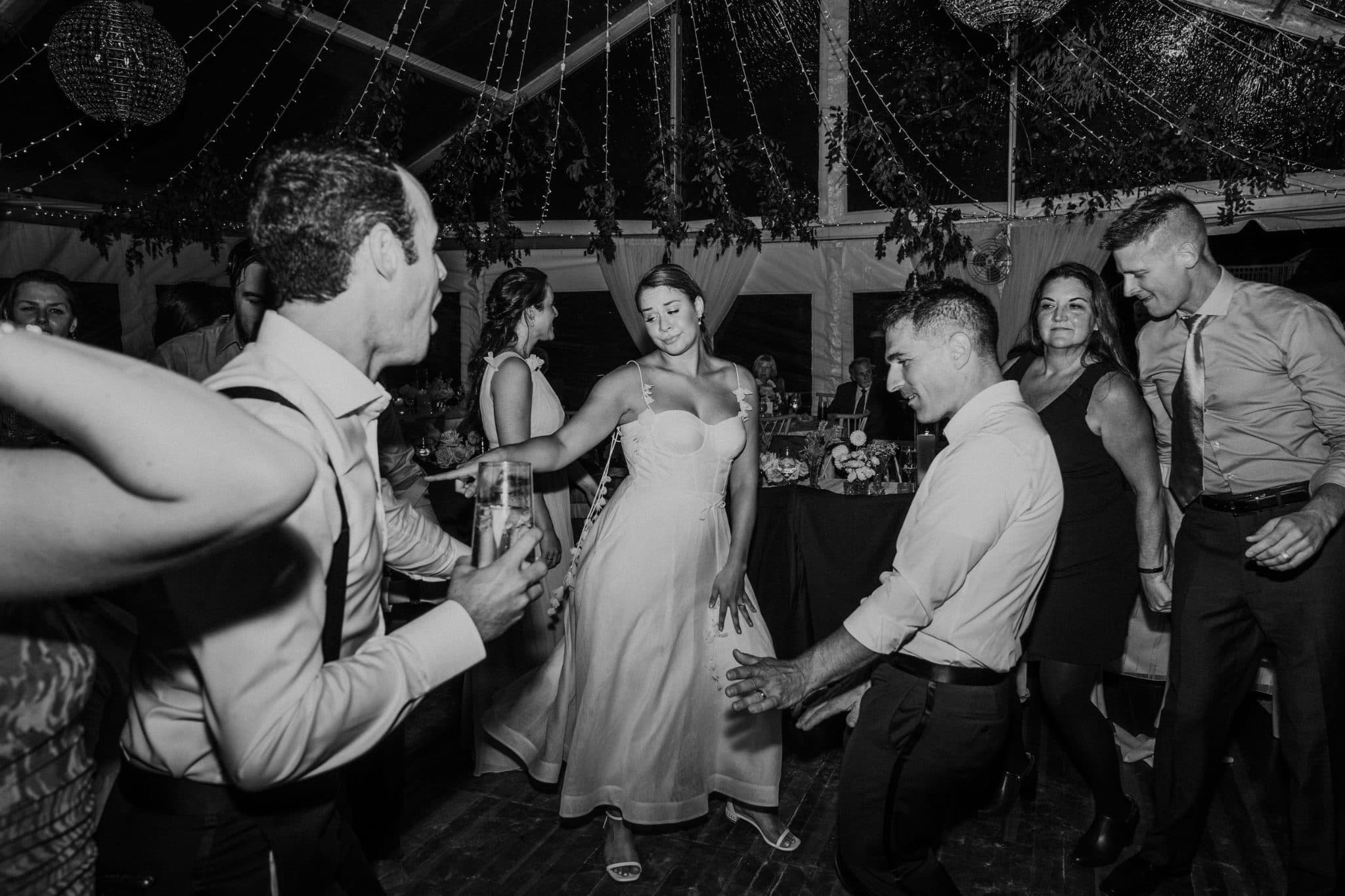 A black-and-white wedding scene shows a bride in a dress dancing with guests under a tent adorned with hanging decorations.