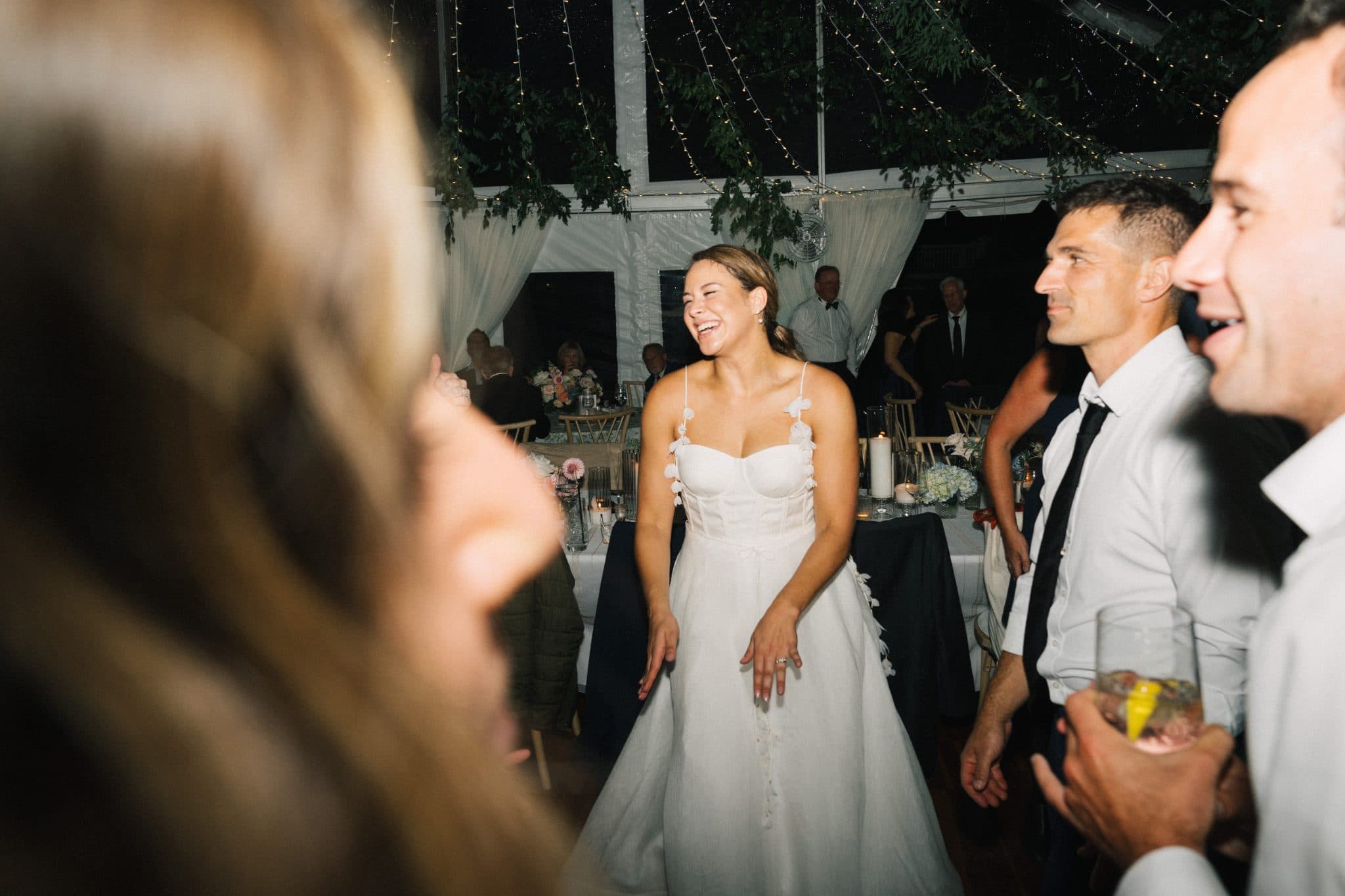 A bride in a white dress laughs while surrounded by guests at a wedding reception under a tent.