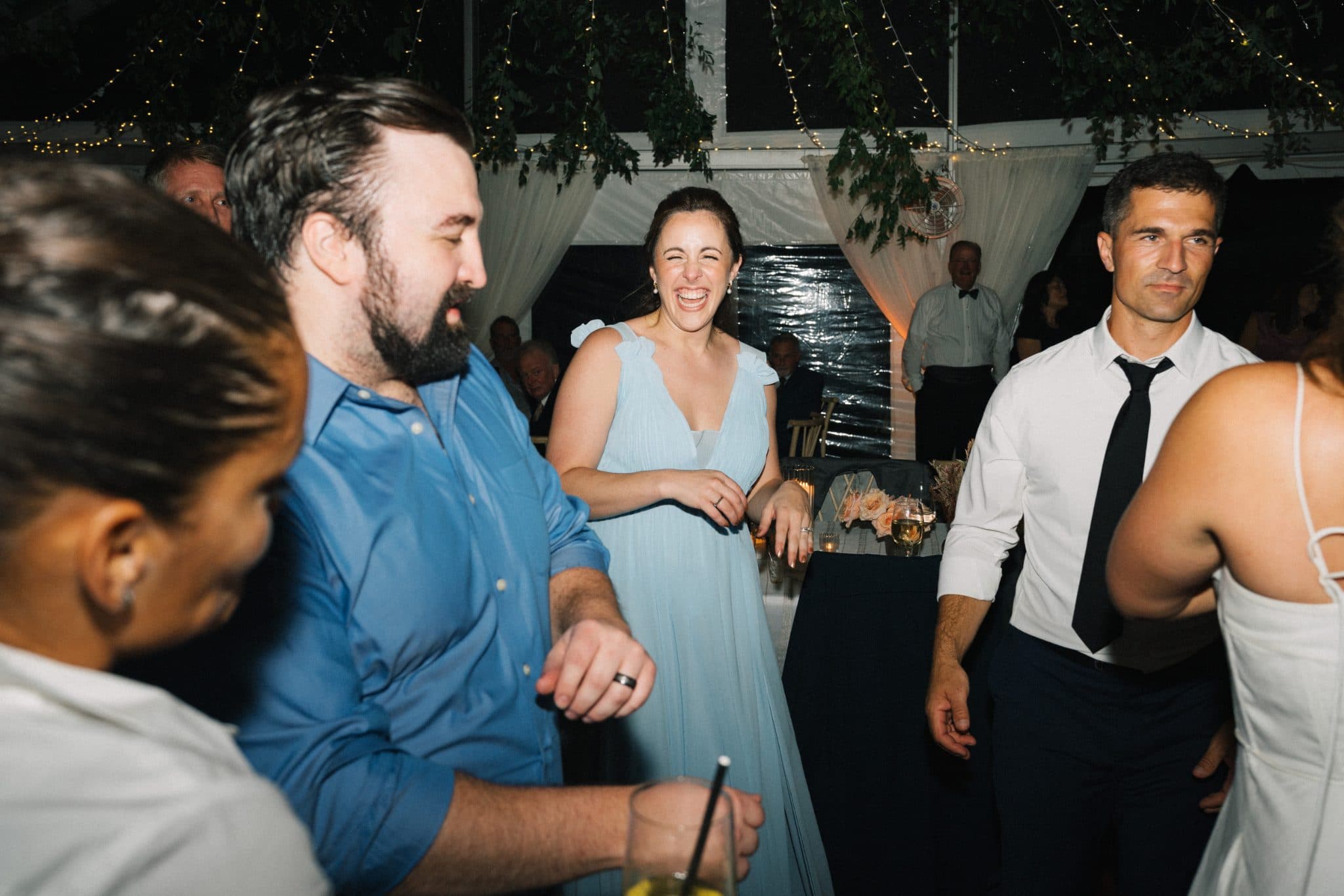 A woman in a blue dress laughs while surrounded by guests at a wedding reception.