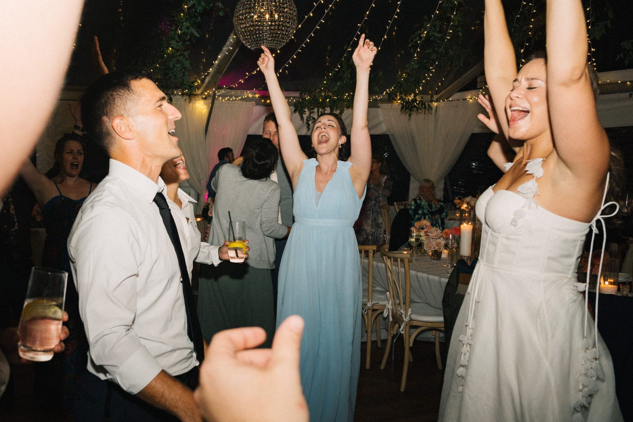 A bride in a white dress dances joyfully with guests in a festive outdoor setting adorned with string lights and a disco ball.
