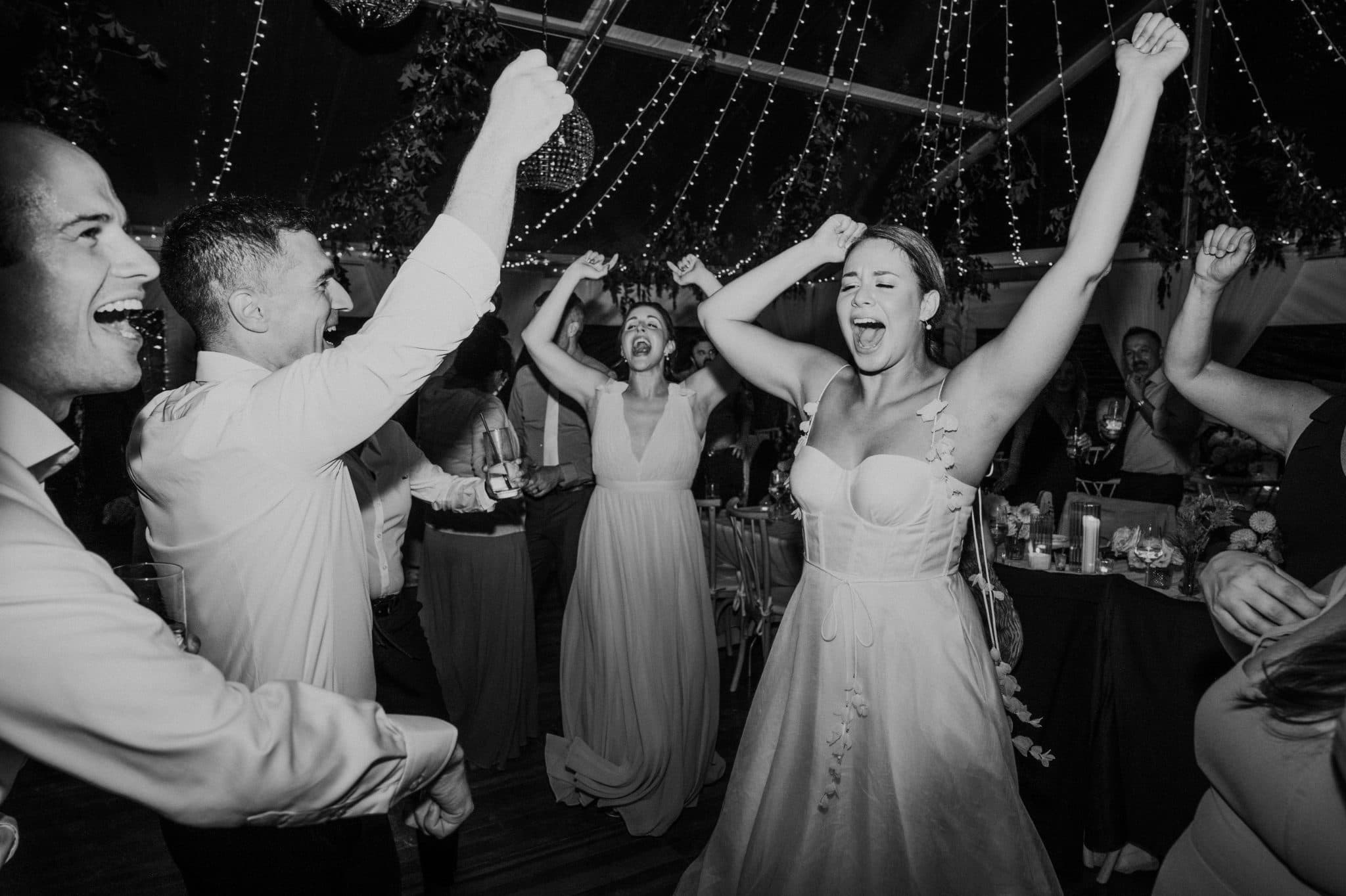 A group of people in formal attire dances joyfully under string lights at a wedding reception.