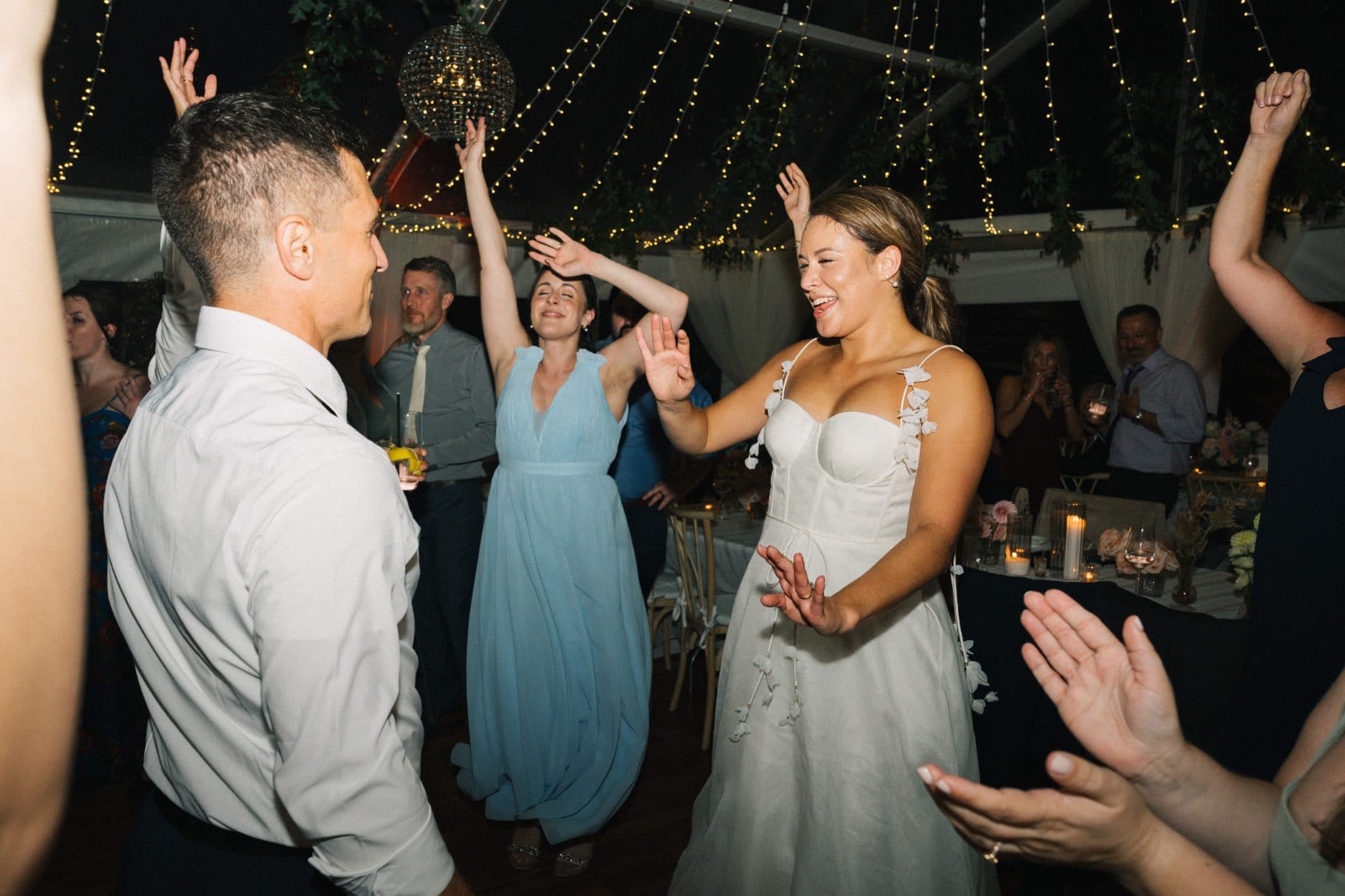 A bride in a white dress dances with guests at a wedding reception under string lights.