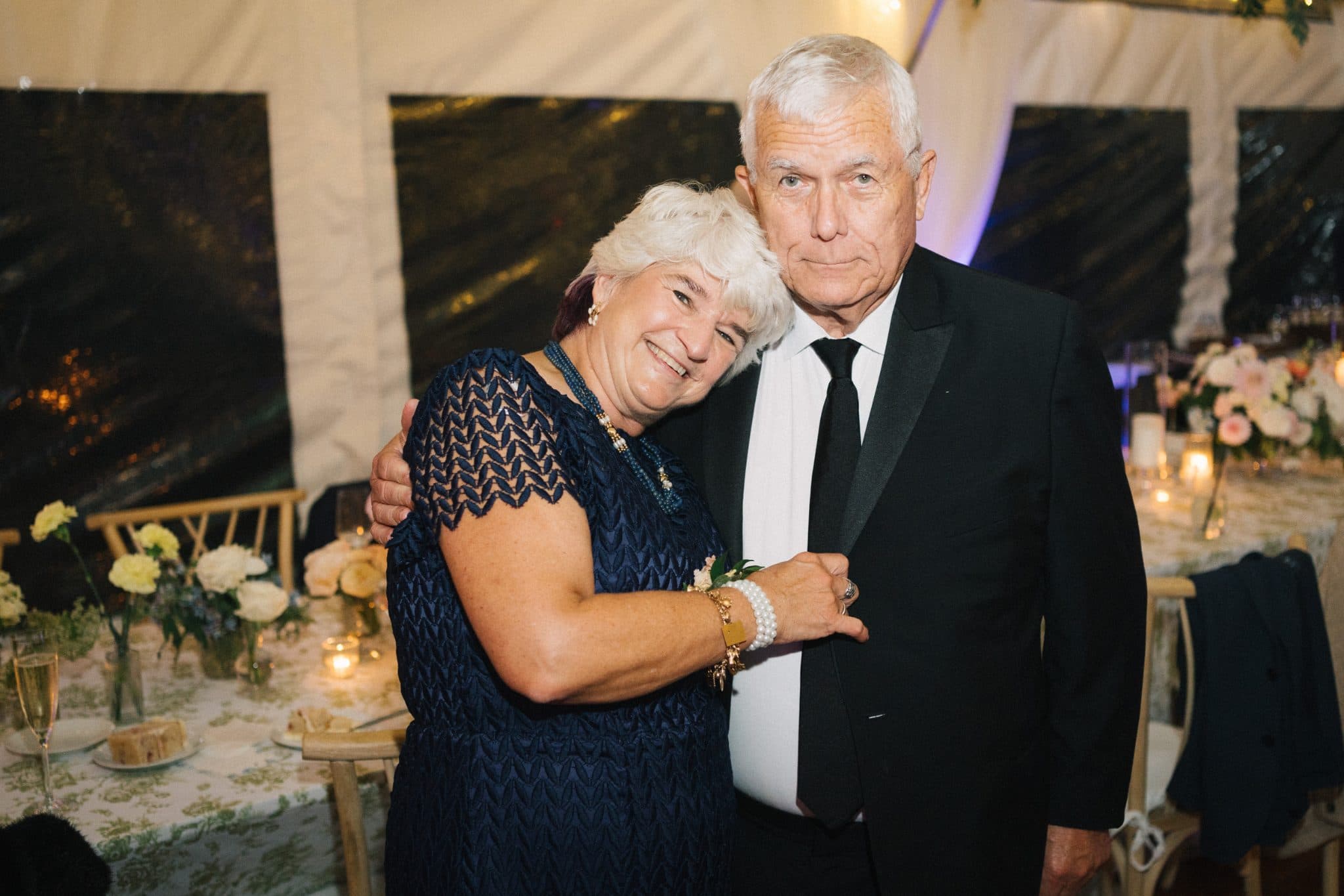 An older couple stands together at a wedding reception, smiling and embracing, with a decorated table and floral arrangements in the background.