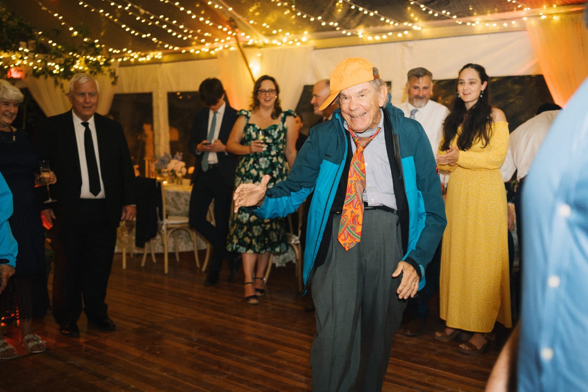 A man in a blue jacket and orange tie dances on a wooden floor surrounded by guests in a warmly lit room.
