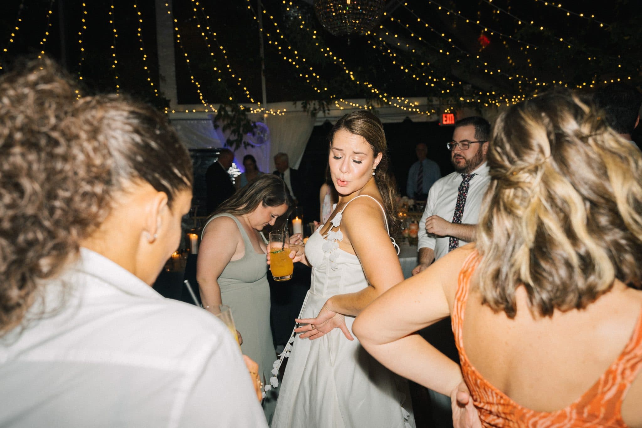 A bride dances among guests at a wedding reception under string lights.