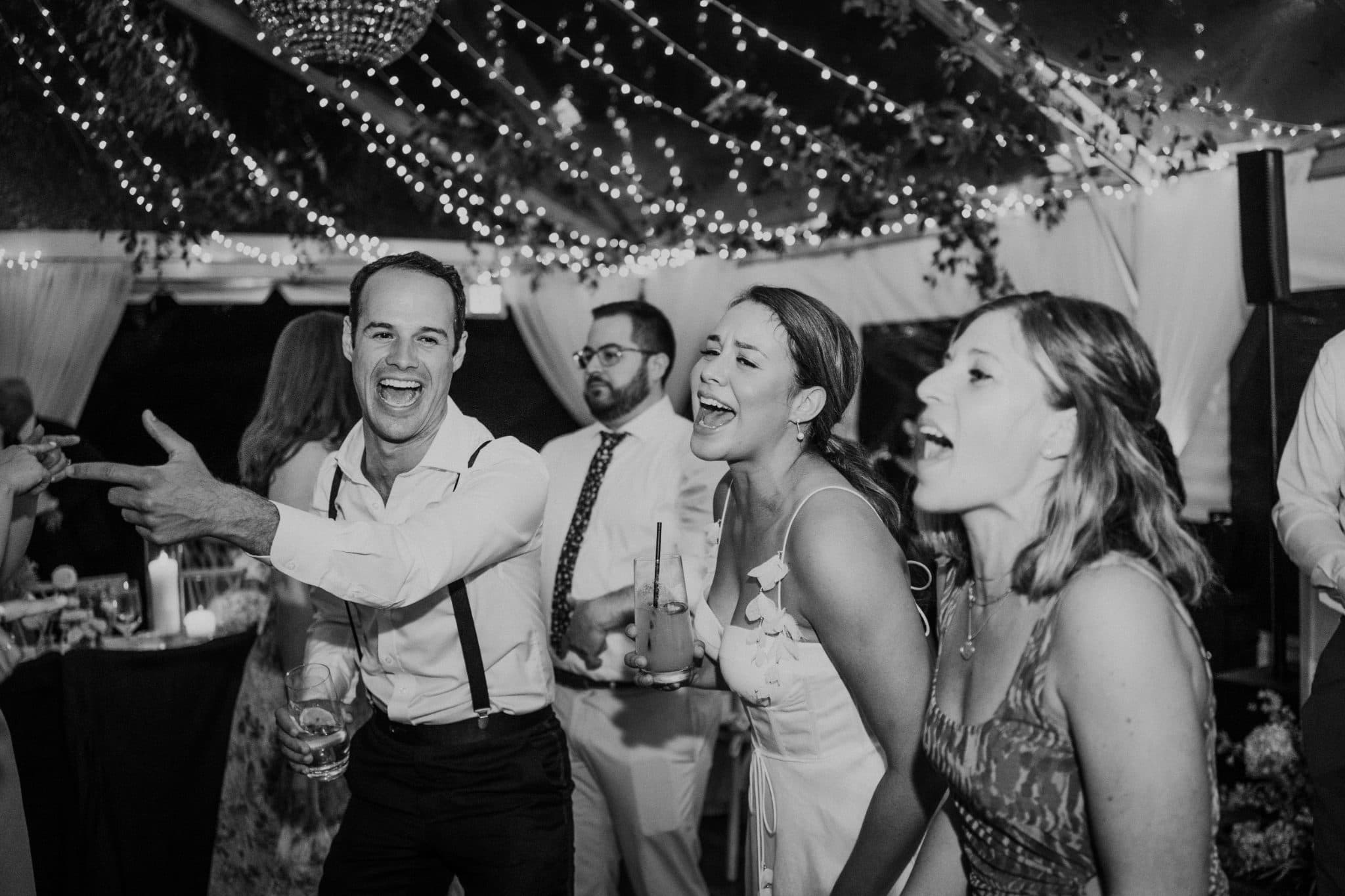 A black-and-white wedding scene shows a man in suspenders dancing, a woman in a white dress laughing, and another woman in a patterned dress singing, under a tent with string lights.