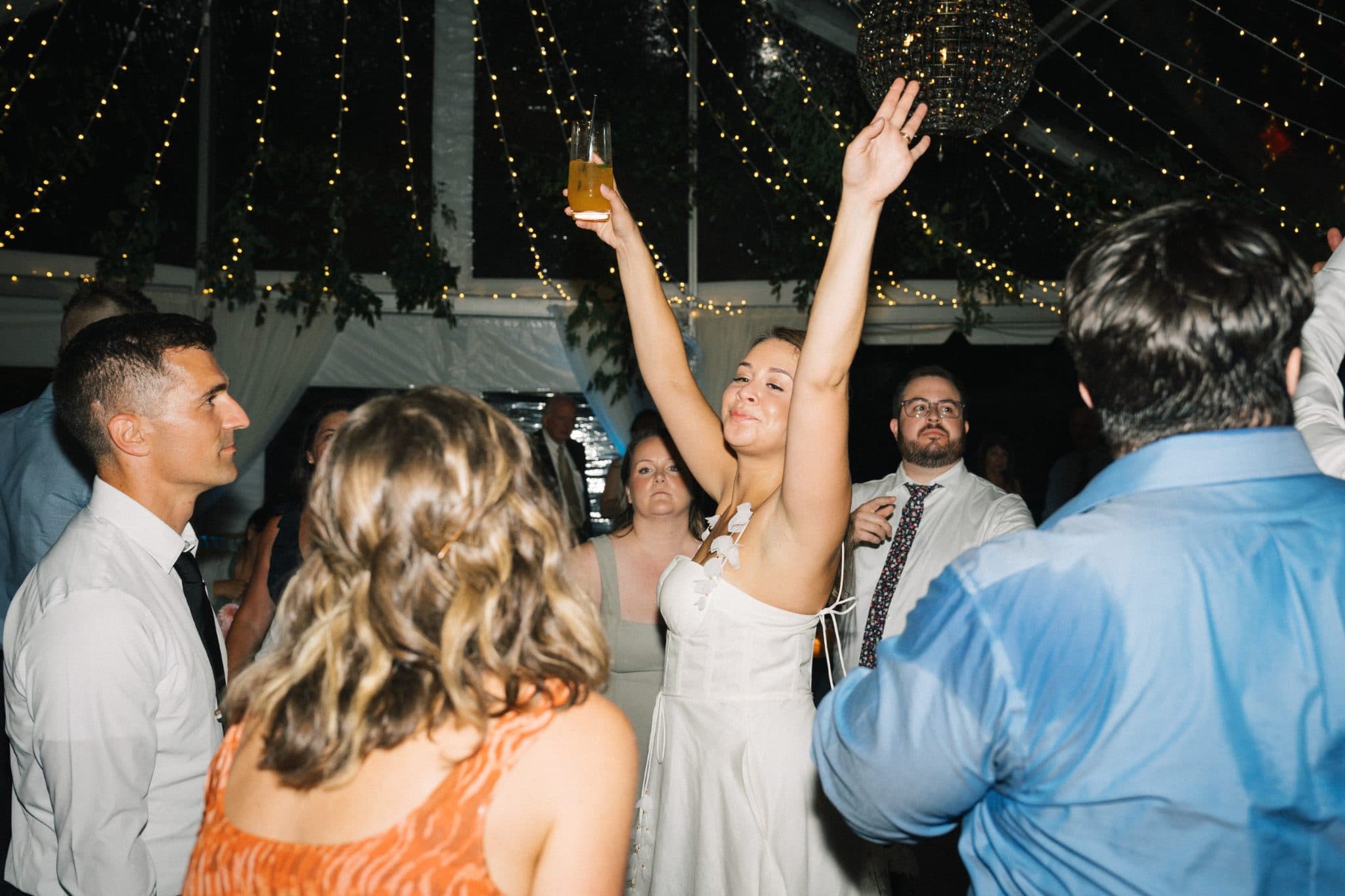 A bride in a white dress raises her arms in celebration at a wedding reception under string lights.