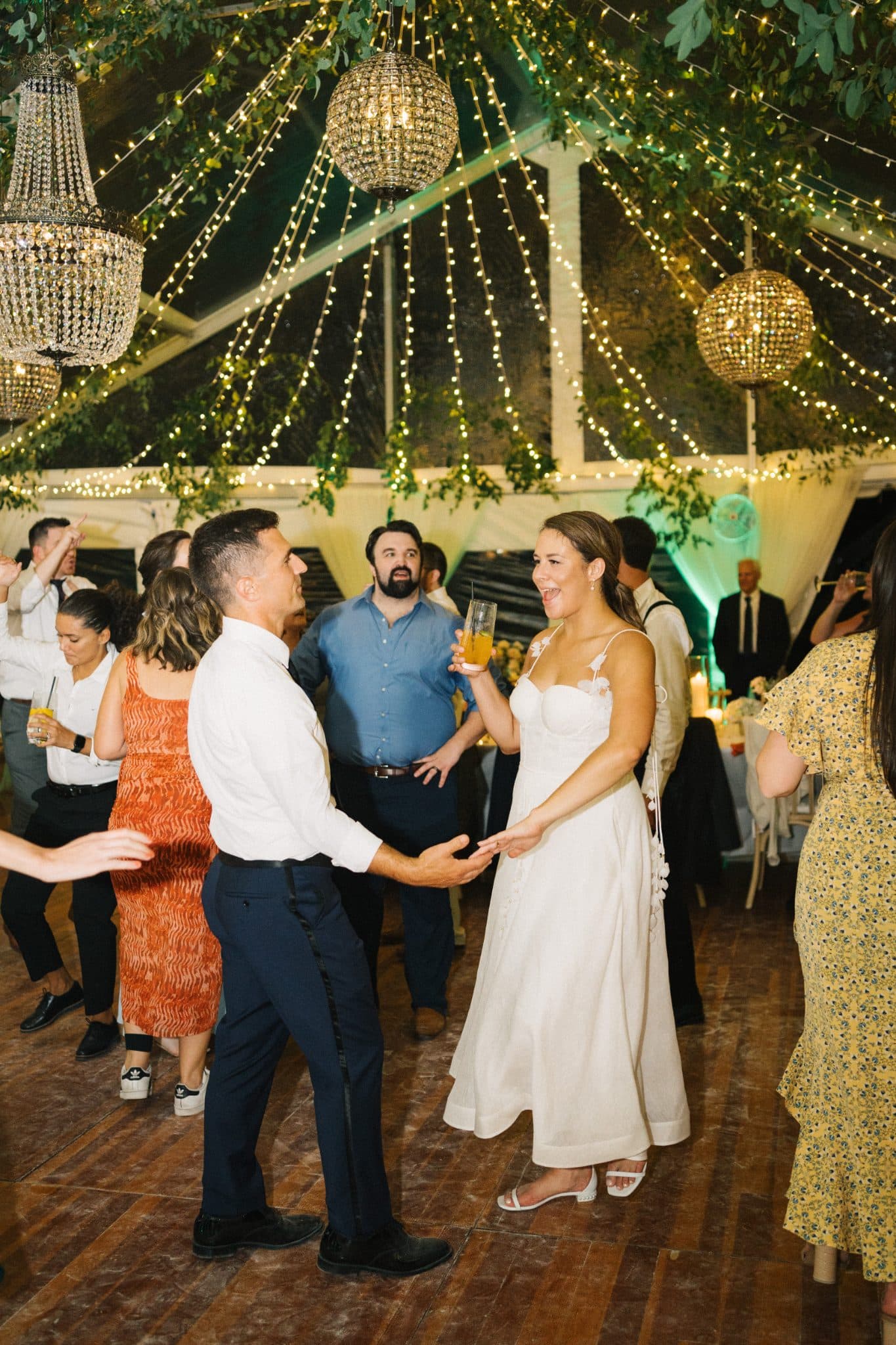 A bride in a white dress dances with a man in a white shirt and dark pants under a festive tent with string lights and chandeliers.