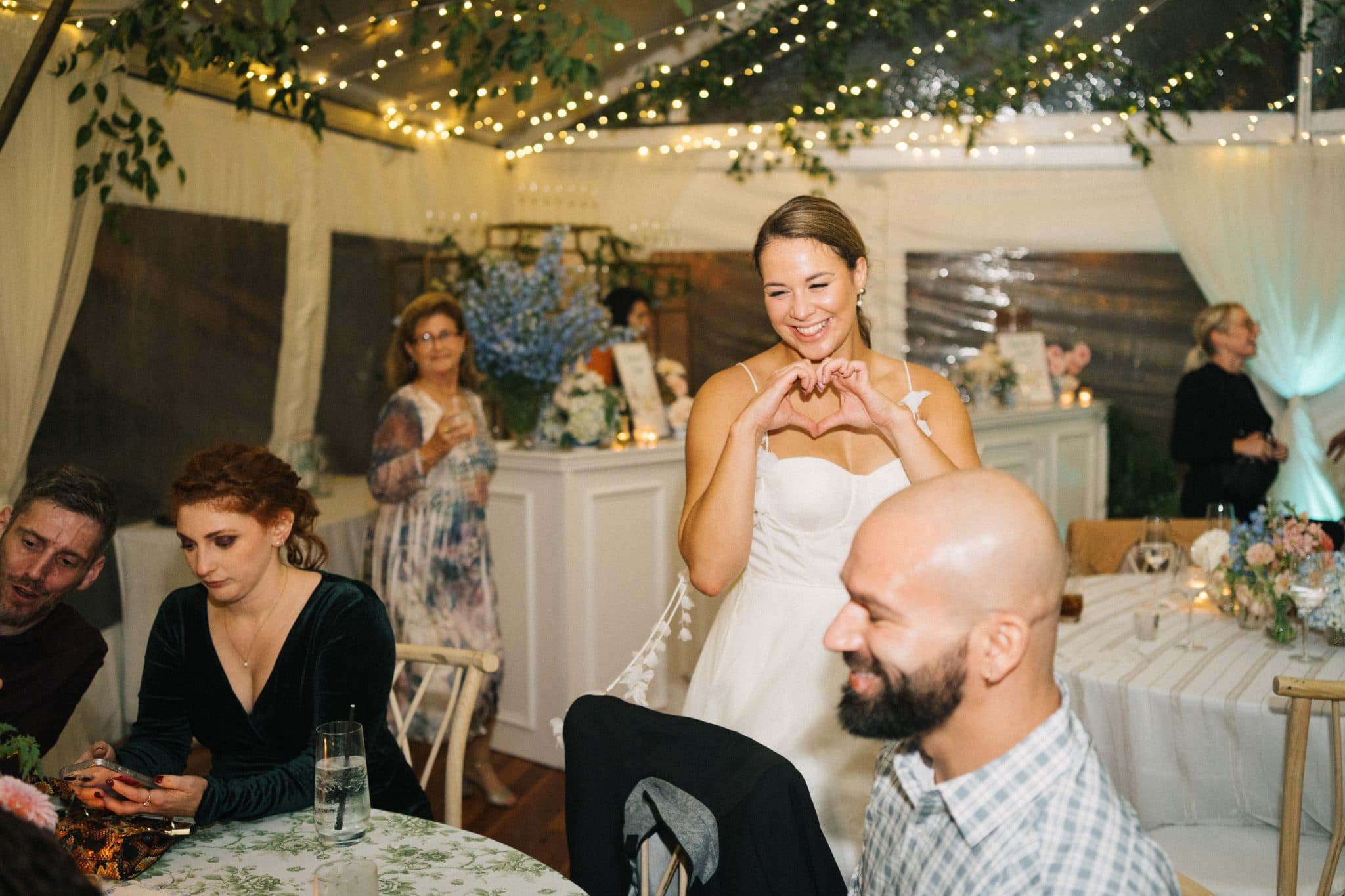 A bride in a white dress makes a heart shape with her hands while seated guests look on at a wedding reception.